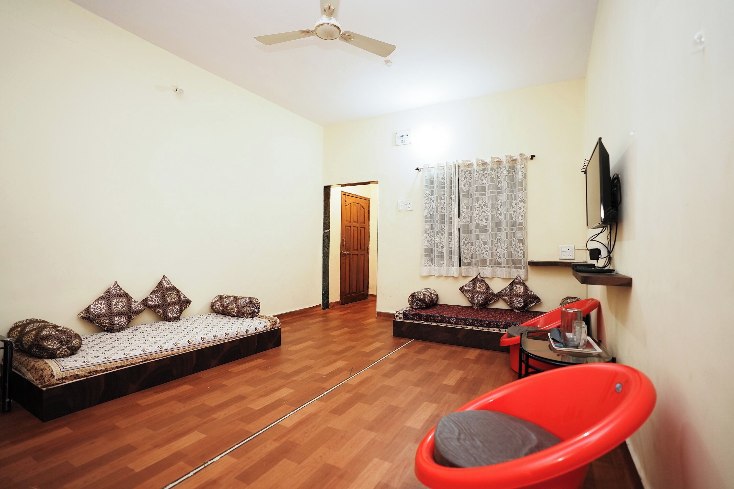 A room with a wooden floor, a ceiling fan, a sofa with patterned cushions, a red chair, and a television mounted on the wall.
