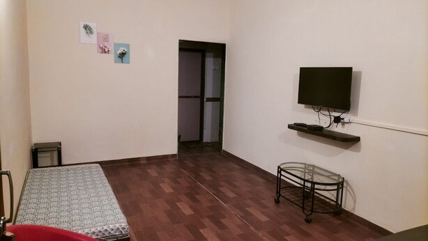 A room with a wooden floor, a bed with a patterned bedspread, a small table, a television mounted on the wall, and a door leading to another room. - Springfield Family Apartment (Mahabaleshwar)