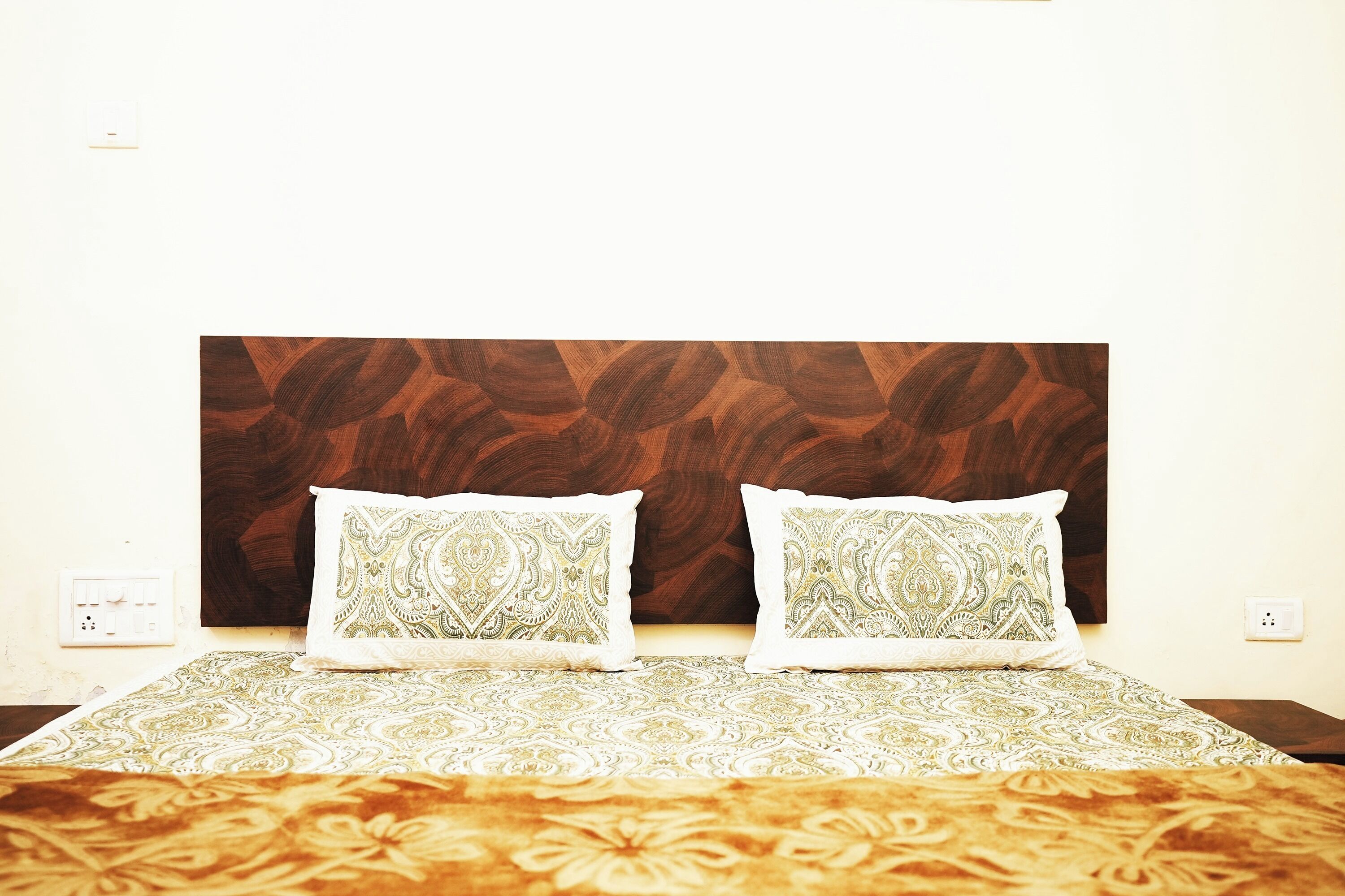 Een bed met beddengoed met patroon en twee decoratieve kussens, een houten hoofdbord en een effen muur met een stopcontact.