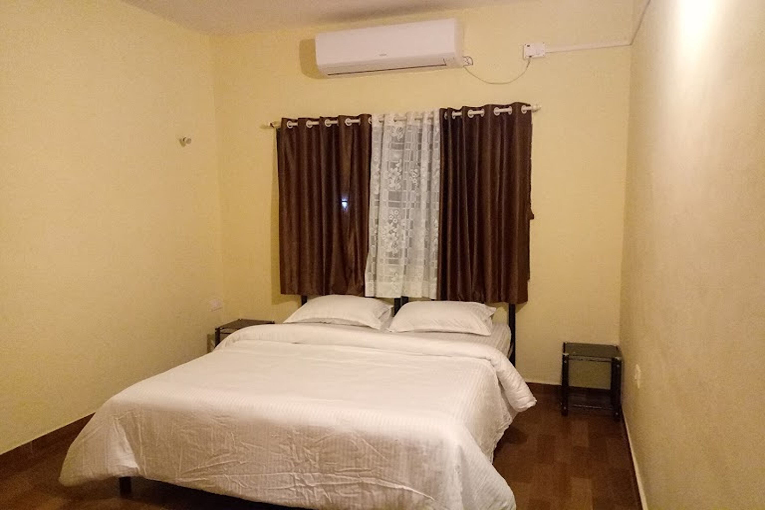 Un dormitorio con cama, cortinas, un equipo de aire acondicionado y una mesita de noche pequeña.