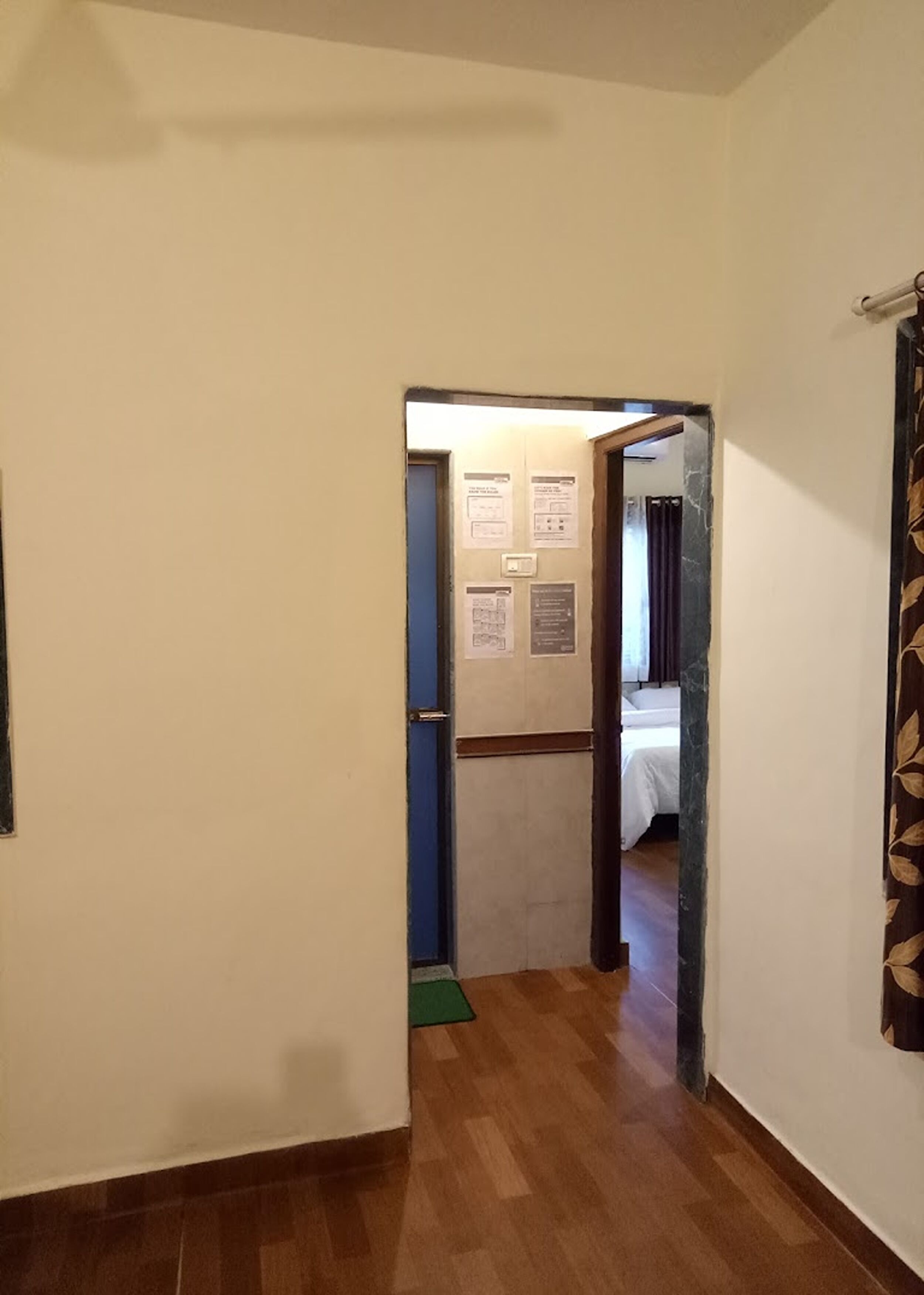 A hallway with a wooden floor leading to a room with a bed and a door with a blue frame. There are several notices on the wall.