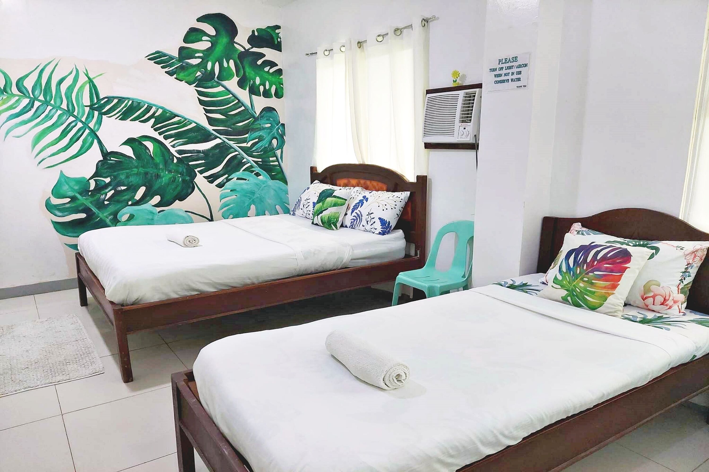 A room with two wooden beds, one with a white bedspread and the other with a white bedspread and a rolled towel. There is a green chair, a large tropical leaf mural on the wall, and a sign on the wall.