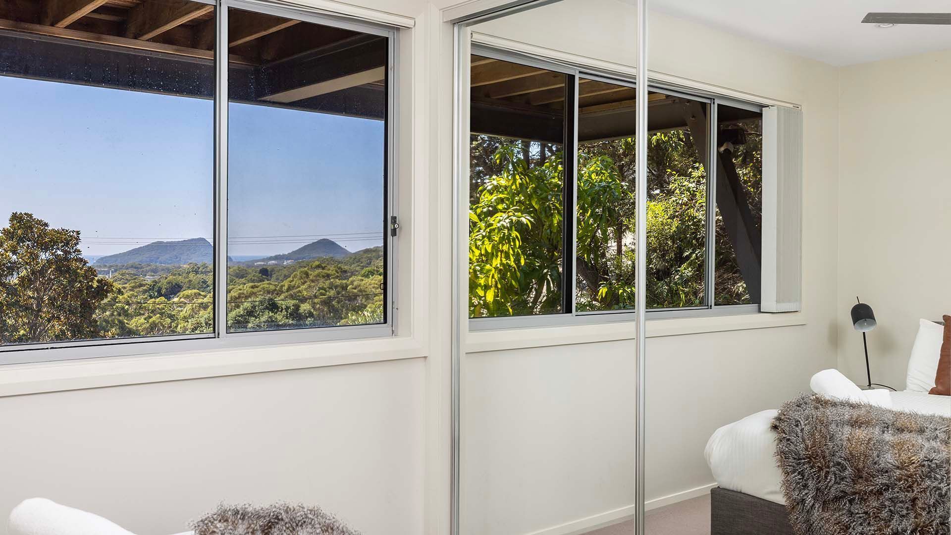 Ein Zimmer mit großen Schiebeglasfenstern, die auf einen Balkon führen und einen Blick auf eine Berglandschaft und viel Grün bieten.