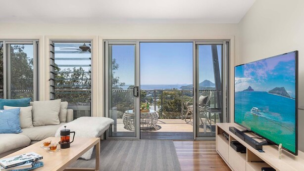 A modern living room with a large TV, comfortable seating, and a balcony with a scenic view.