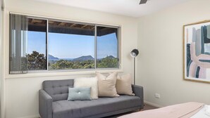 A room with a grey sofa, a large window with a view of greenery and hills, and a floor lamp.