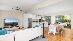 A modern living room with a sofa, a dining area with a table and chairs, a kitchen with white cabinets, and a large window offering a view of greenery.