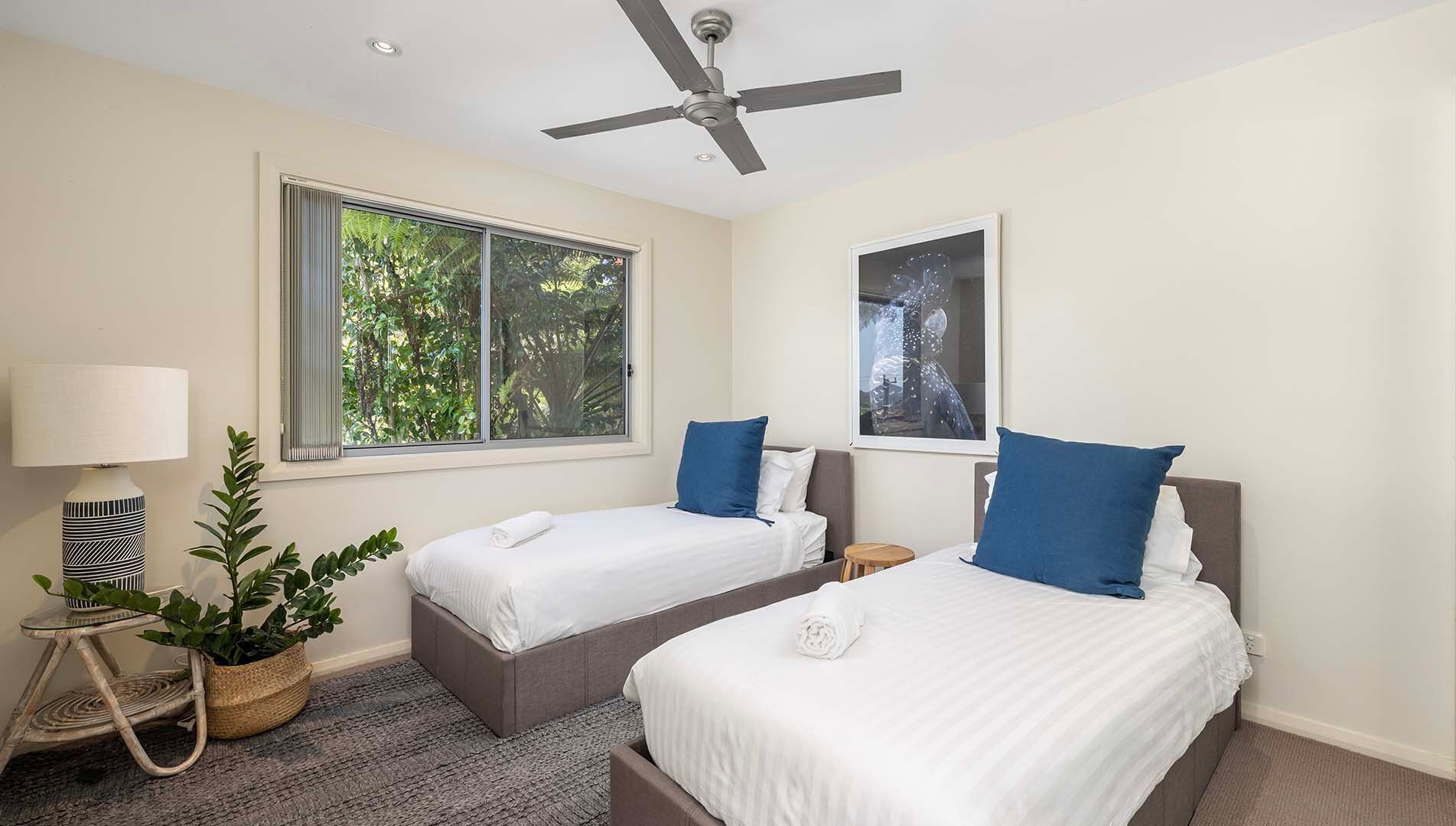 Une chambre d’hôtel avec deux lits, un ventilateur de plafond, une fenêtre donnant sur des arbres, une lampe et une plante en pot.