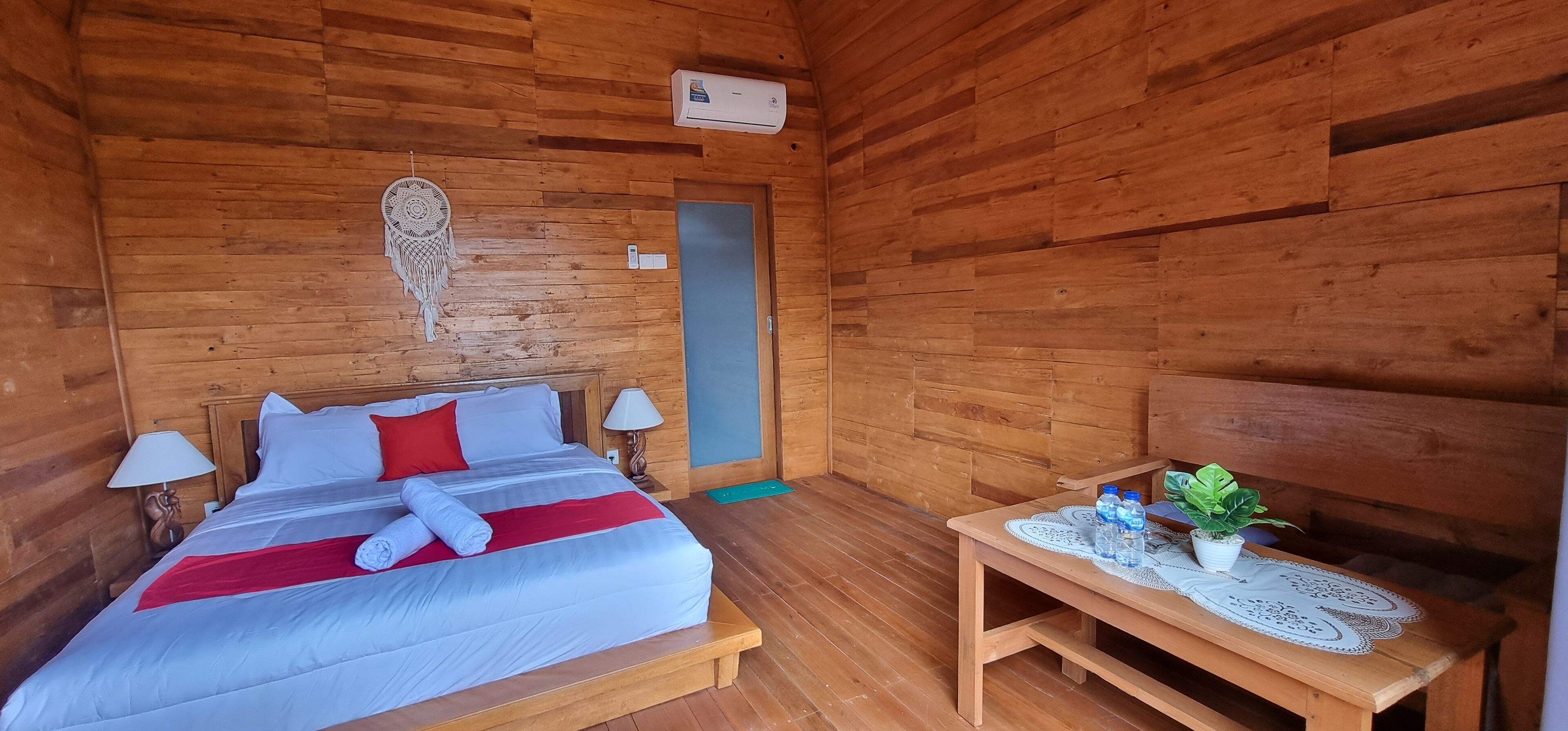 Una habitación de cabaña de madera con cama, lámparas de noche, una mesita con una planta y un atrapasueños en la pared.