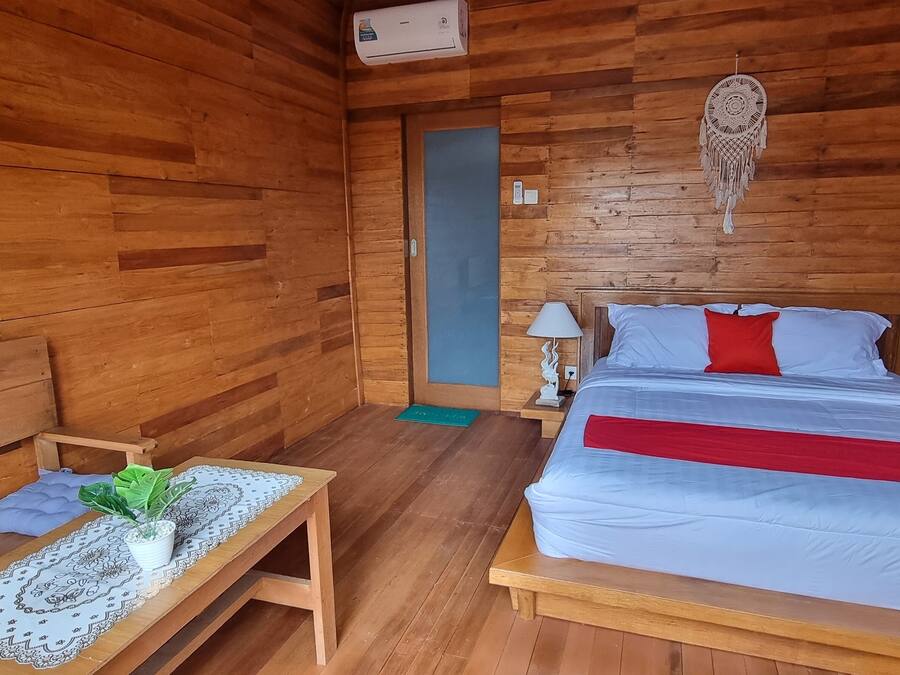 Una habitación de madera con una cama, mesitas de noche, una pequeña mesa con una planta y un atrapasueños en la pared.