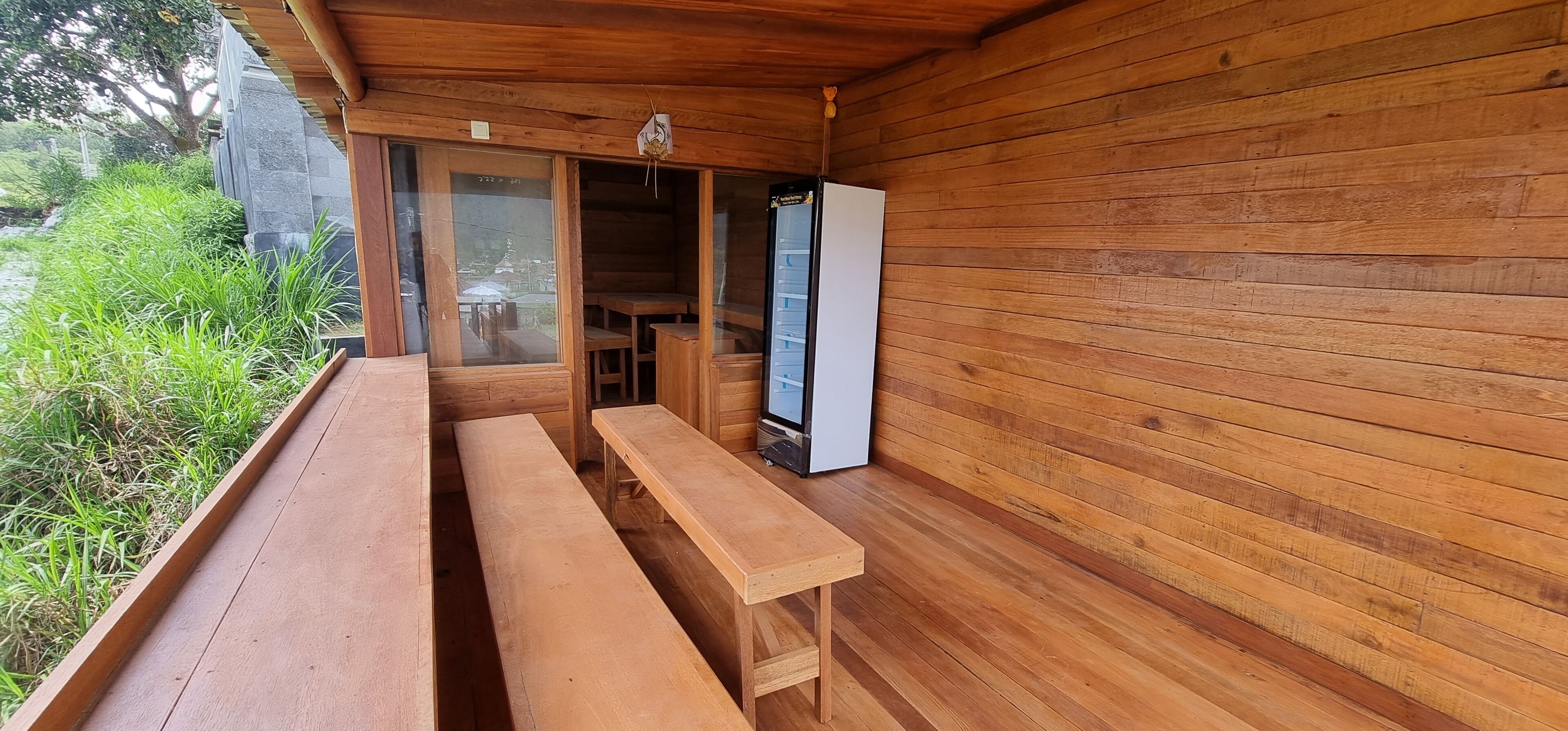 A wooden deck with benches and a refrigerator, surrounded by wooden walls and a view of greenery.