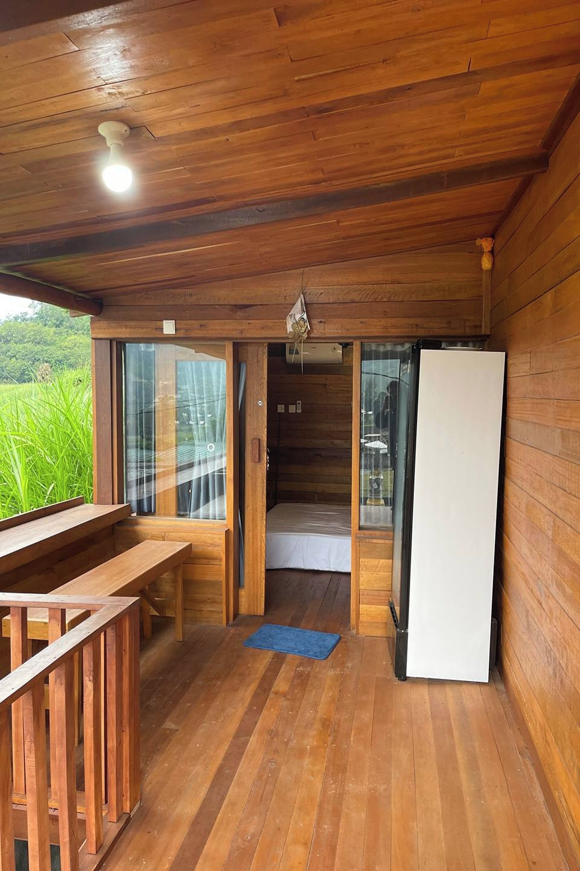 A wooden cabin with a wooden deck, a bed, a refrigerator, and a window overlooking greenery.