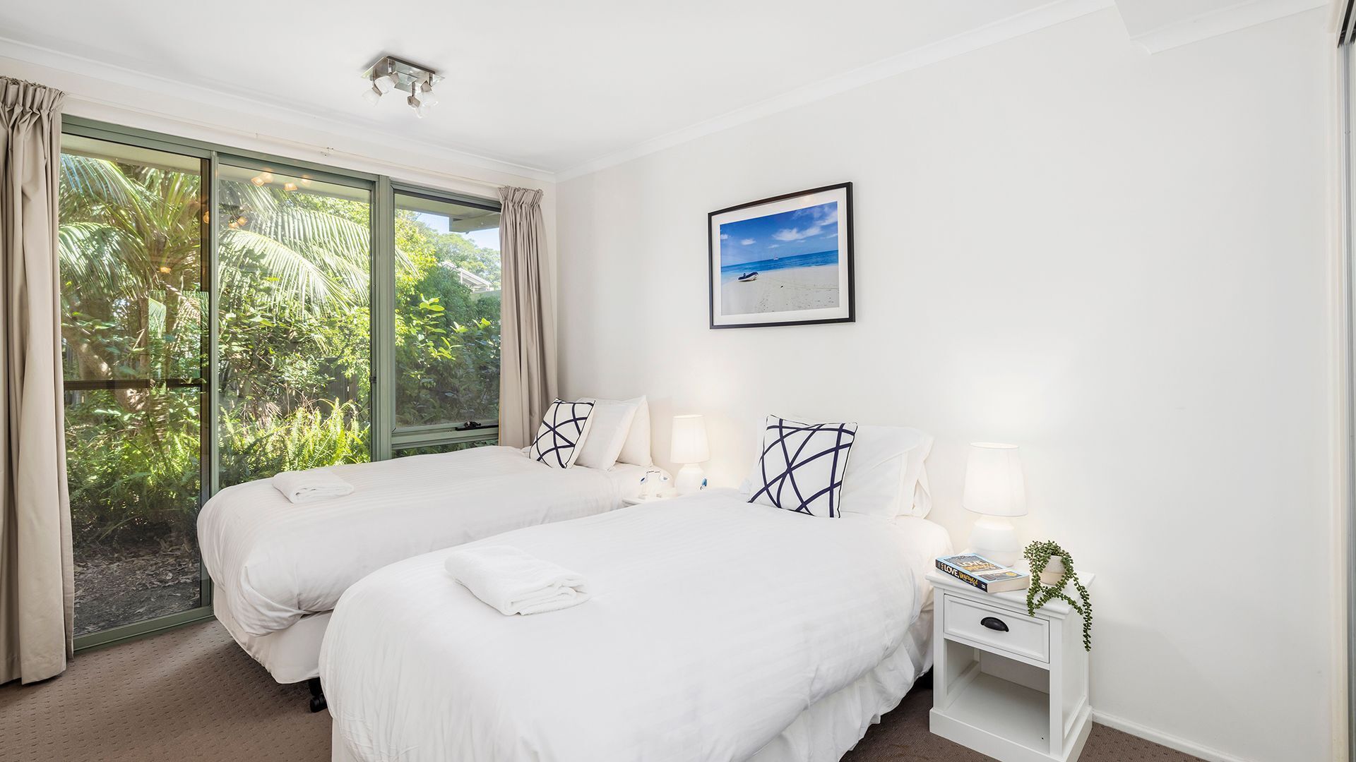 A hotel room with two beds, a large window with curtains, a framed picture on the wall, and a bedside table with a lamp and a book.