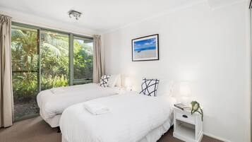 A hotel room with two beds, a large window with curtains, a framed picture on the wall, and a bedside table with a lamp and a book.