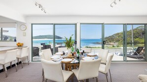 Un espace repas offrant une vue sur l’océan, avec une table ronde dressée pour six personnes, des chaises blanches et une grande fenêtre donnant sur la plage et l’océan.
