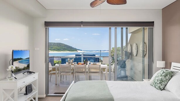 Ein Schlafzimmer mit einem großen Bett, einem Fernseher auf einem Schreibtisch und einem Balkon mit Blick auf den Strand.