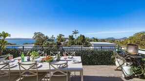 Un espace extérieur aménagé pour les repas, avec vue sur l’océan et les collines, une table dressée pour deux personnes et un barbecue à proximité.