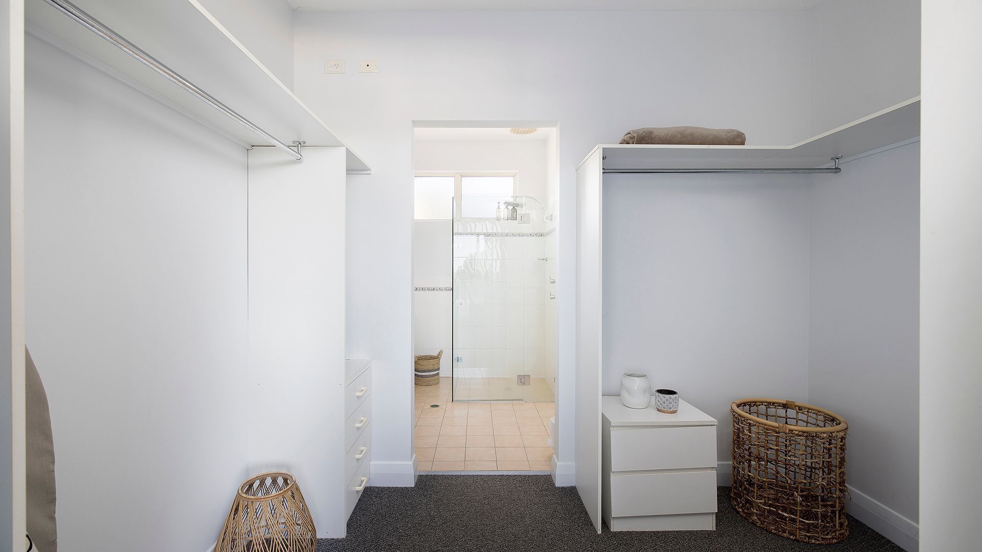 A narrow hallway with a glass shower door, a small white bedside table with a cup, and a wicker basket.