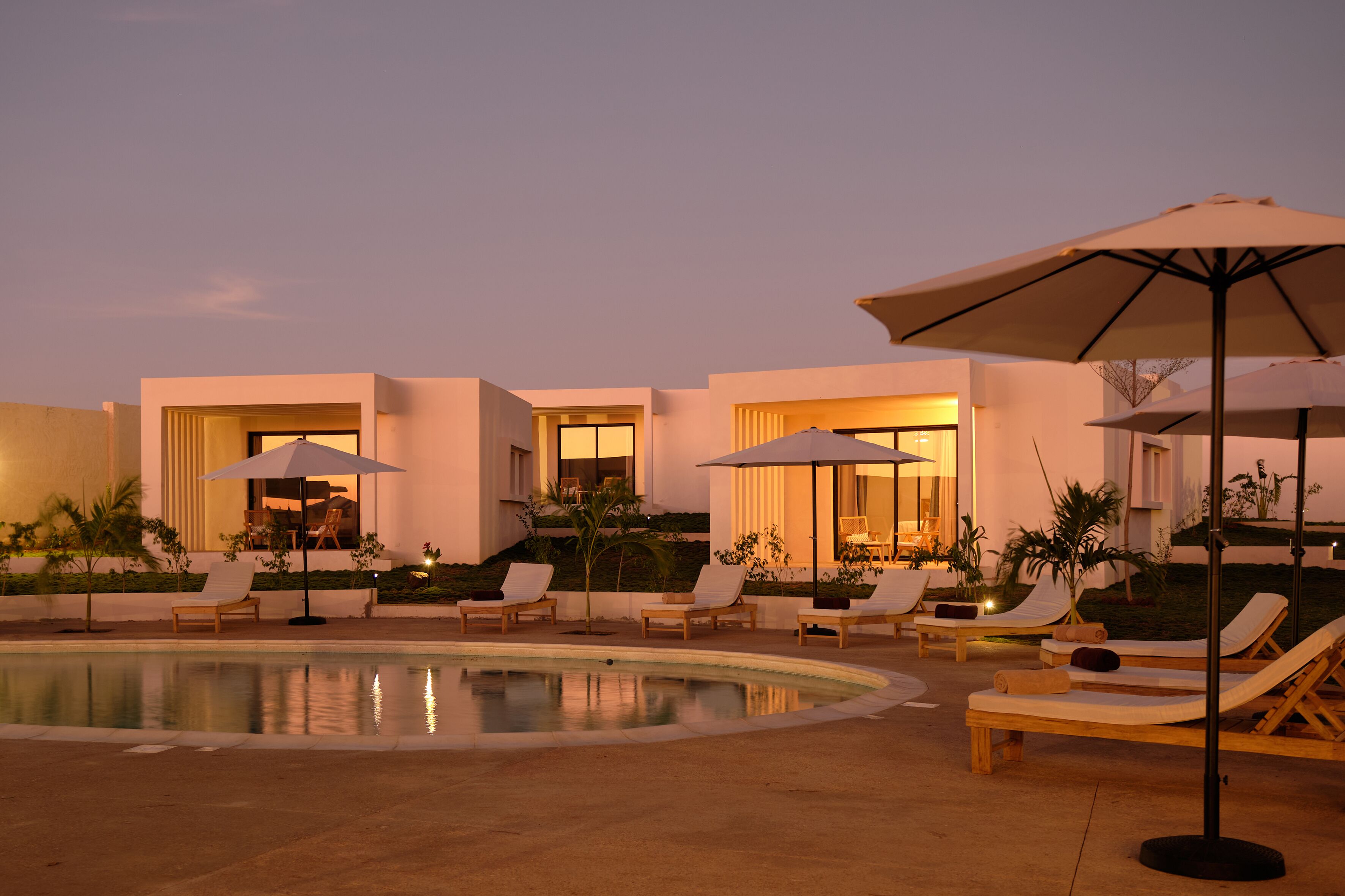 A poolside area with lounge chairs and umbrellas, a pool, and a building with large windows and doors.