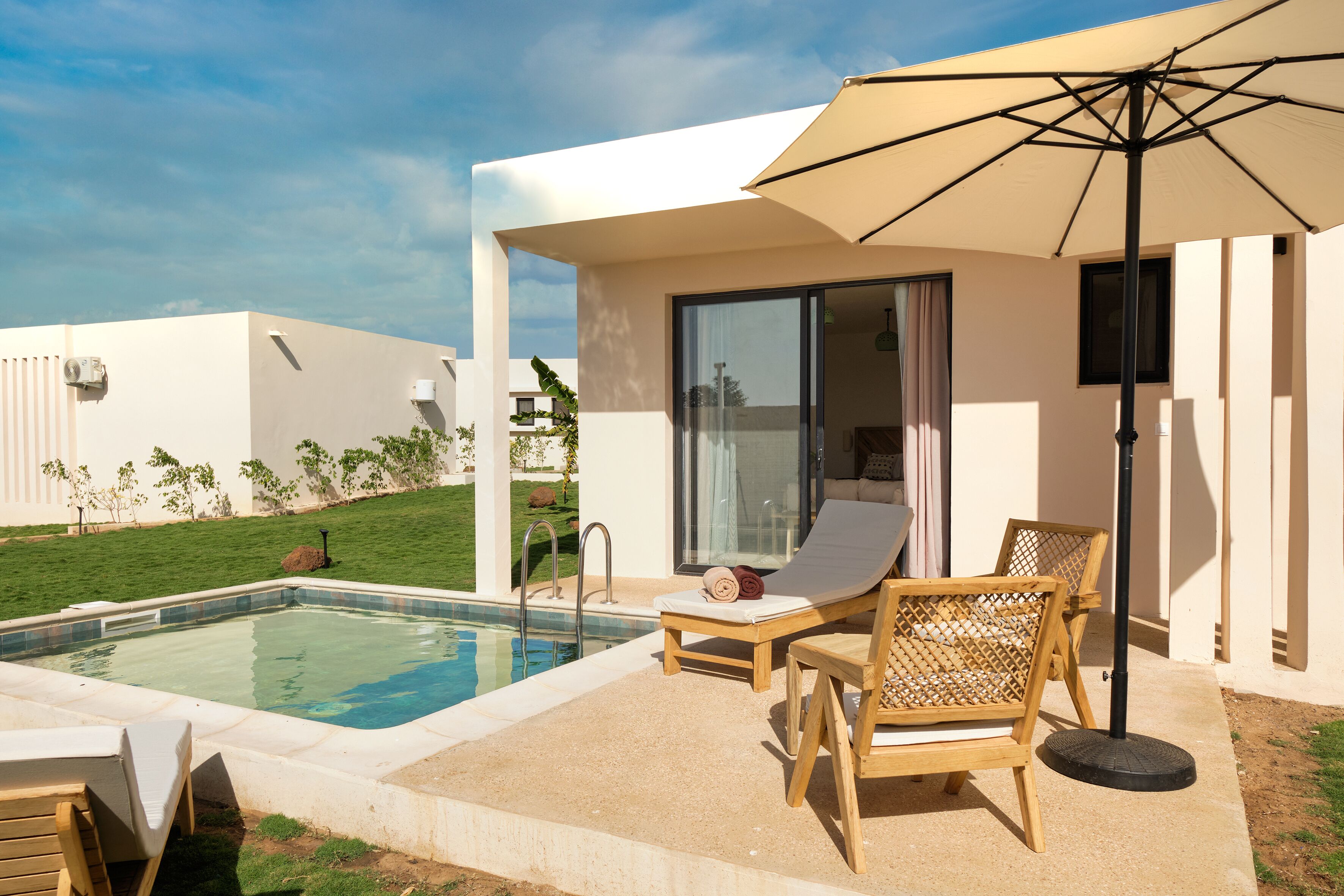 Un espace piscine extérieur moderne, doté de chaises longues, d’un grand parasol et d’une porte vitrée donnant sur un patio abrité.