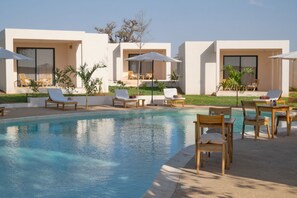 Un área al lado de la piscina con sillones y sombrillas, una piscina azul cristalina y edificios modernos con grandes ventanales.