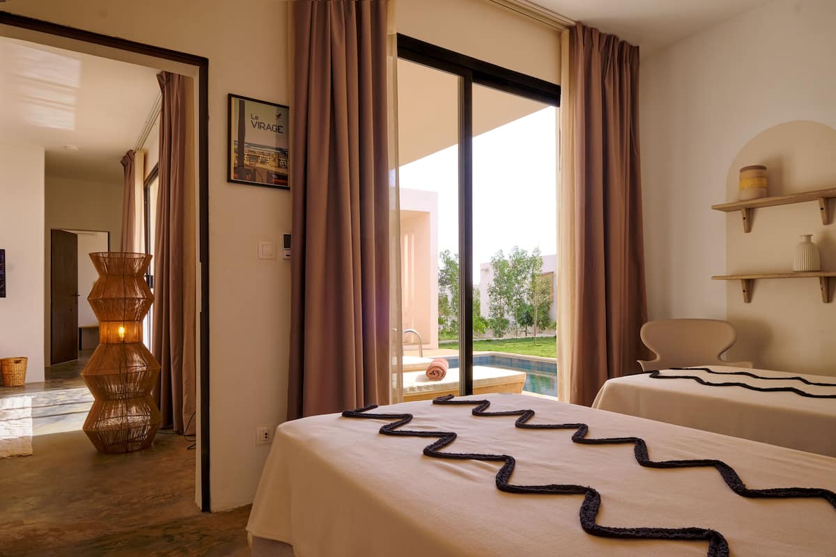 A hotel room with two beds, a large window with curtains, a wicker floor lamp, and a view of a pool and greenery outside.