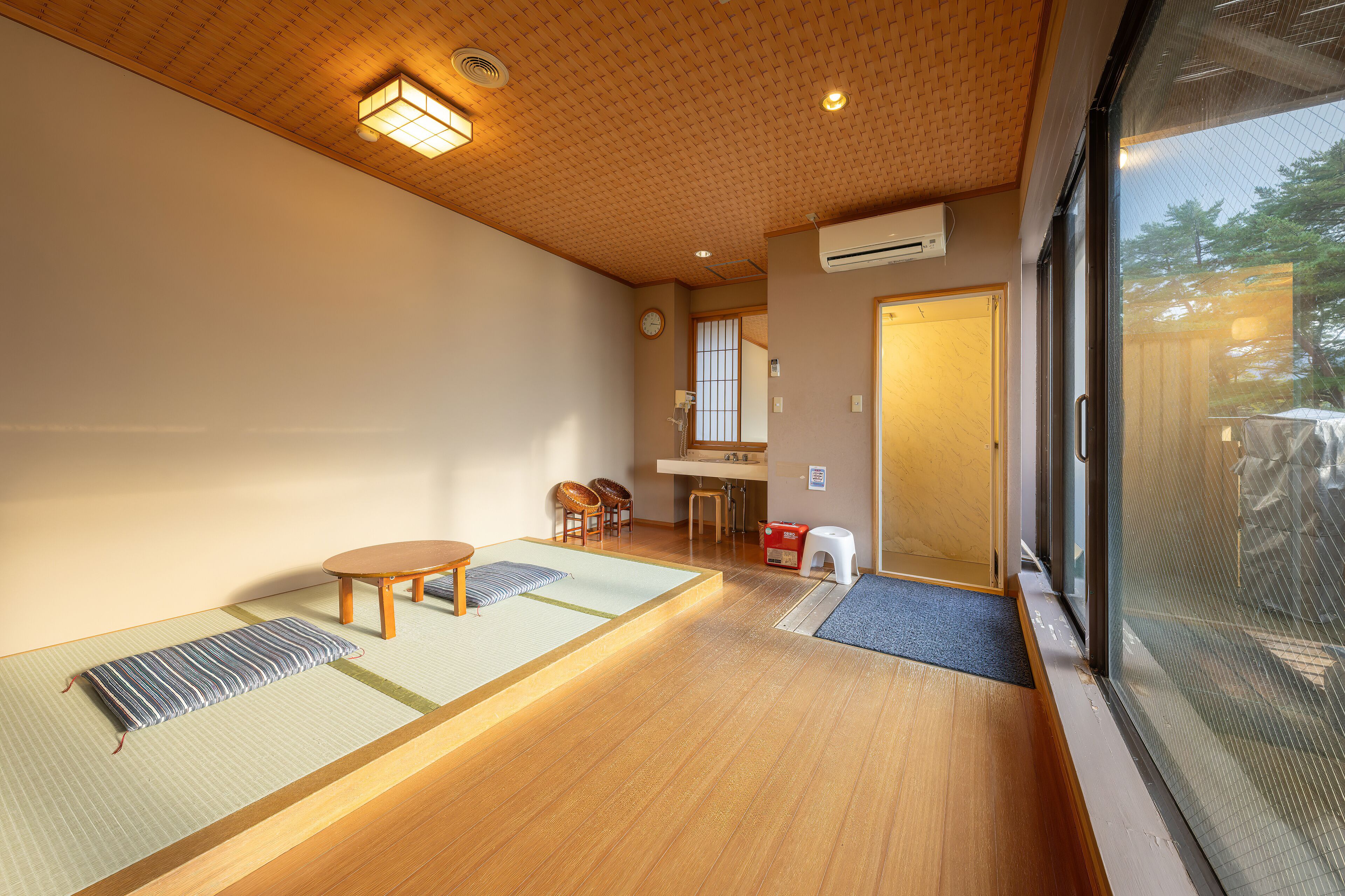 A traditional tatami mat room with a wooden ceiling, a tatami mat table, a low wooden stool, and a sliding door leading to another room.