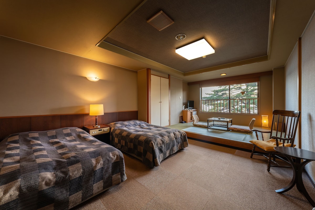 A hotel room with two beds, a desk, a chair, a small table, a window with a view, and a lamp.