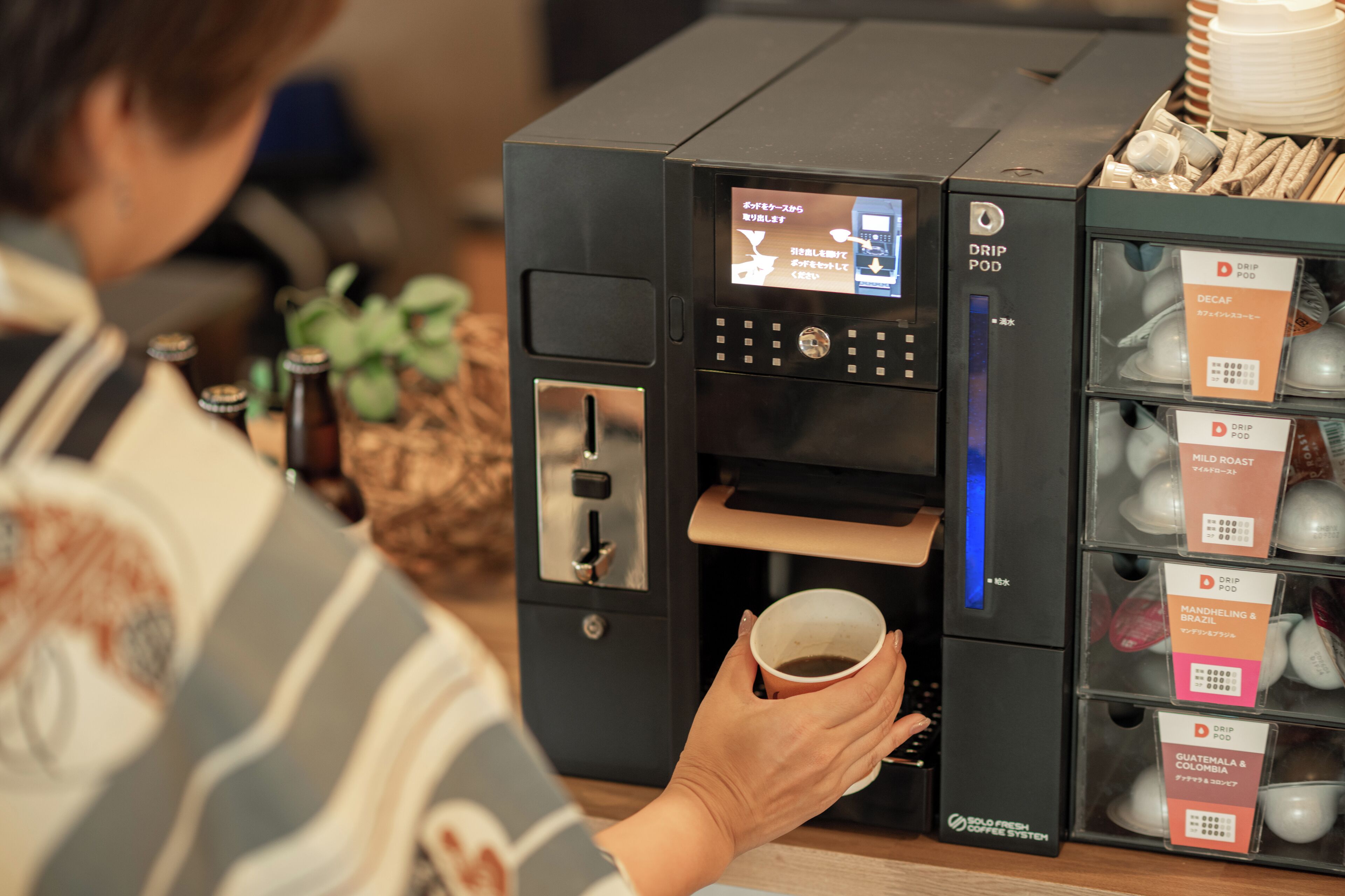 Una cafetera con sistema de cápsulas, una taza de café y cápsulas etiquetadas para distintos tipos de café.
