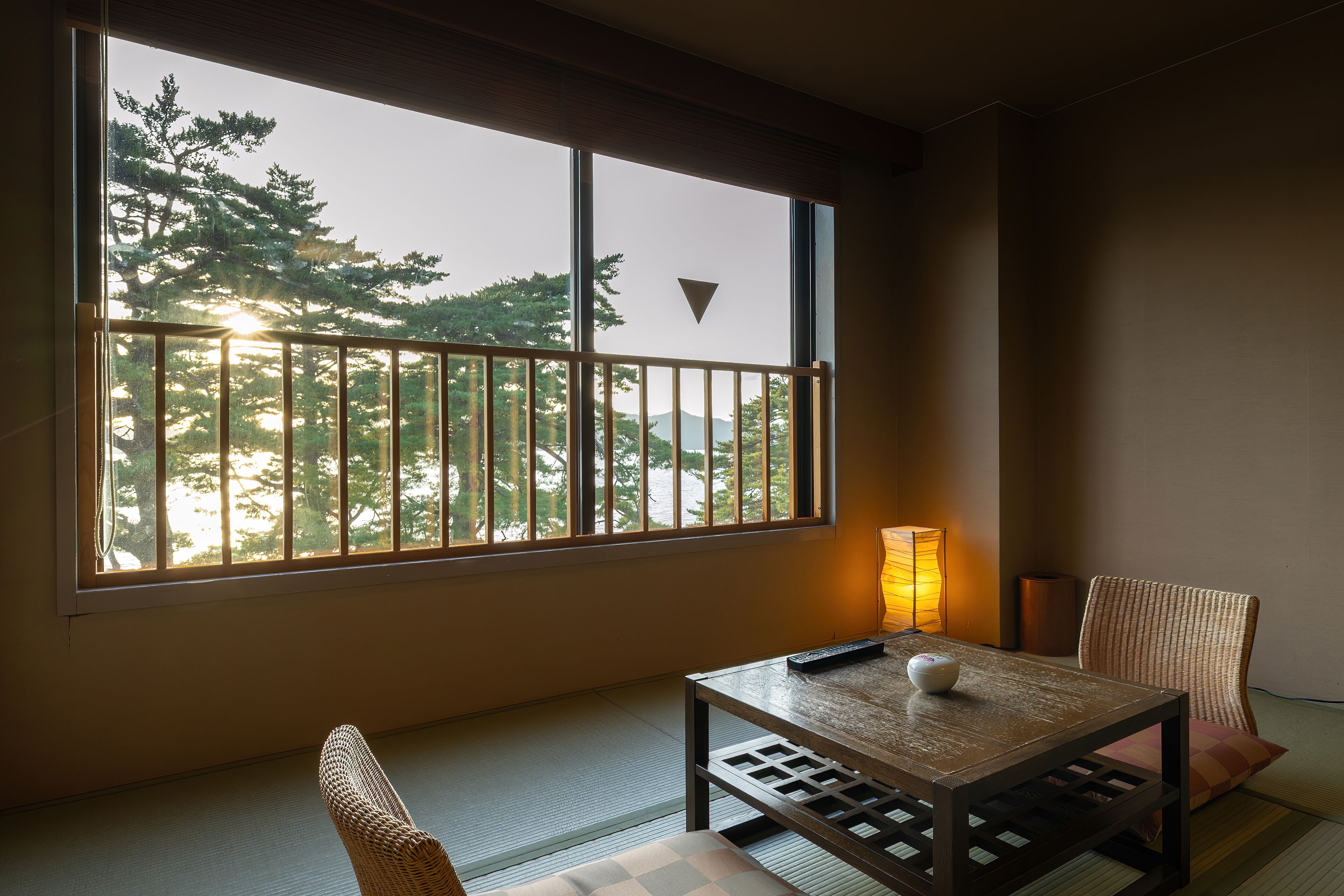 A room with tatami flooring, a low table with a remote control and a decorative item, a chair, and a large window with a view of trees and mountains.