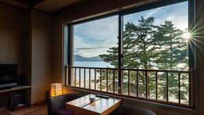 Habitación con una ventana grande que ofrece vistas a árboles y una masa de agua. Hay una mesa con un control remoto y un televisor sobre un soporte a la izquierda.