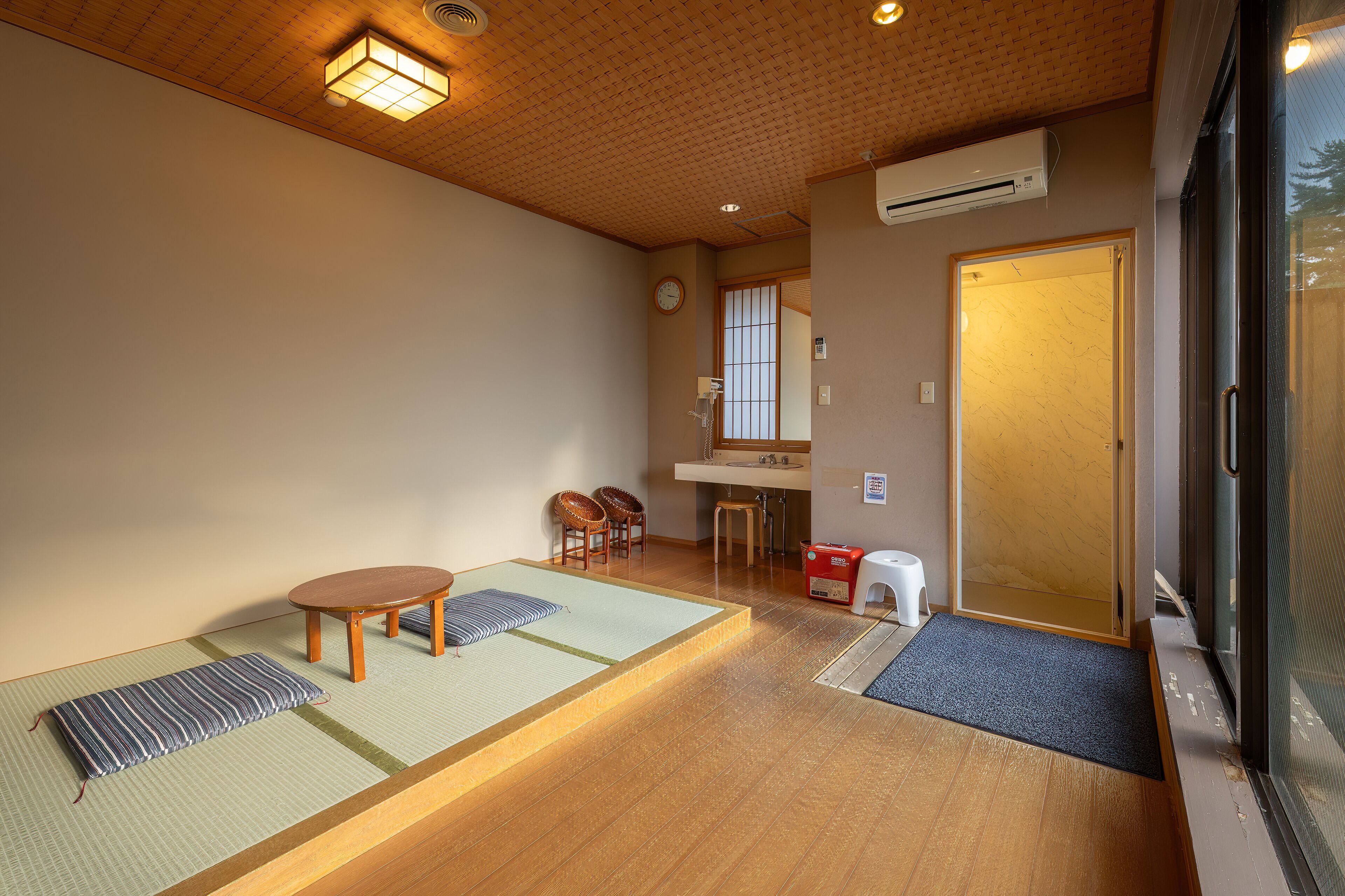 Una habitación japonesa tradicional con esteras de tatami, una mesa de madera, dos sillas, un lavamanos pequeño y una puerta corredera.