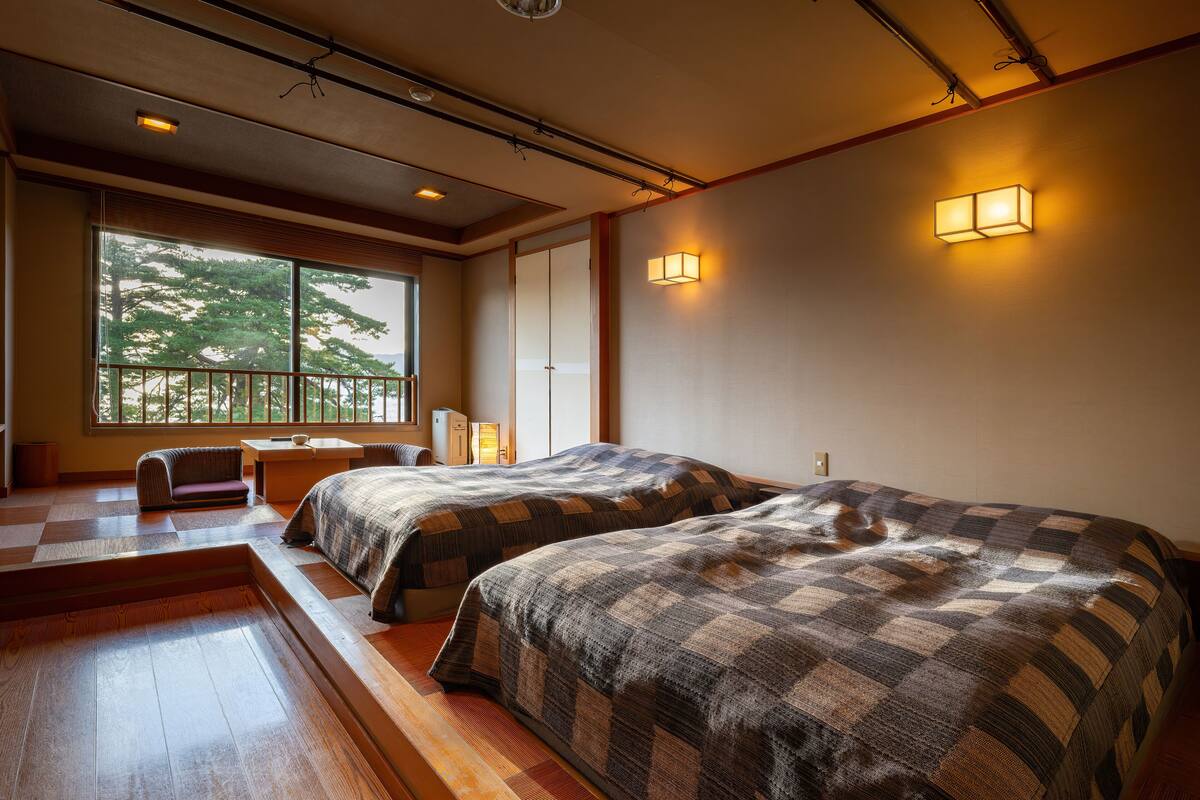 A hotel room with two beds, a small seating area, and a large window offering a view of trees.