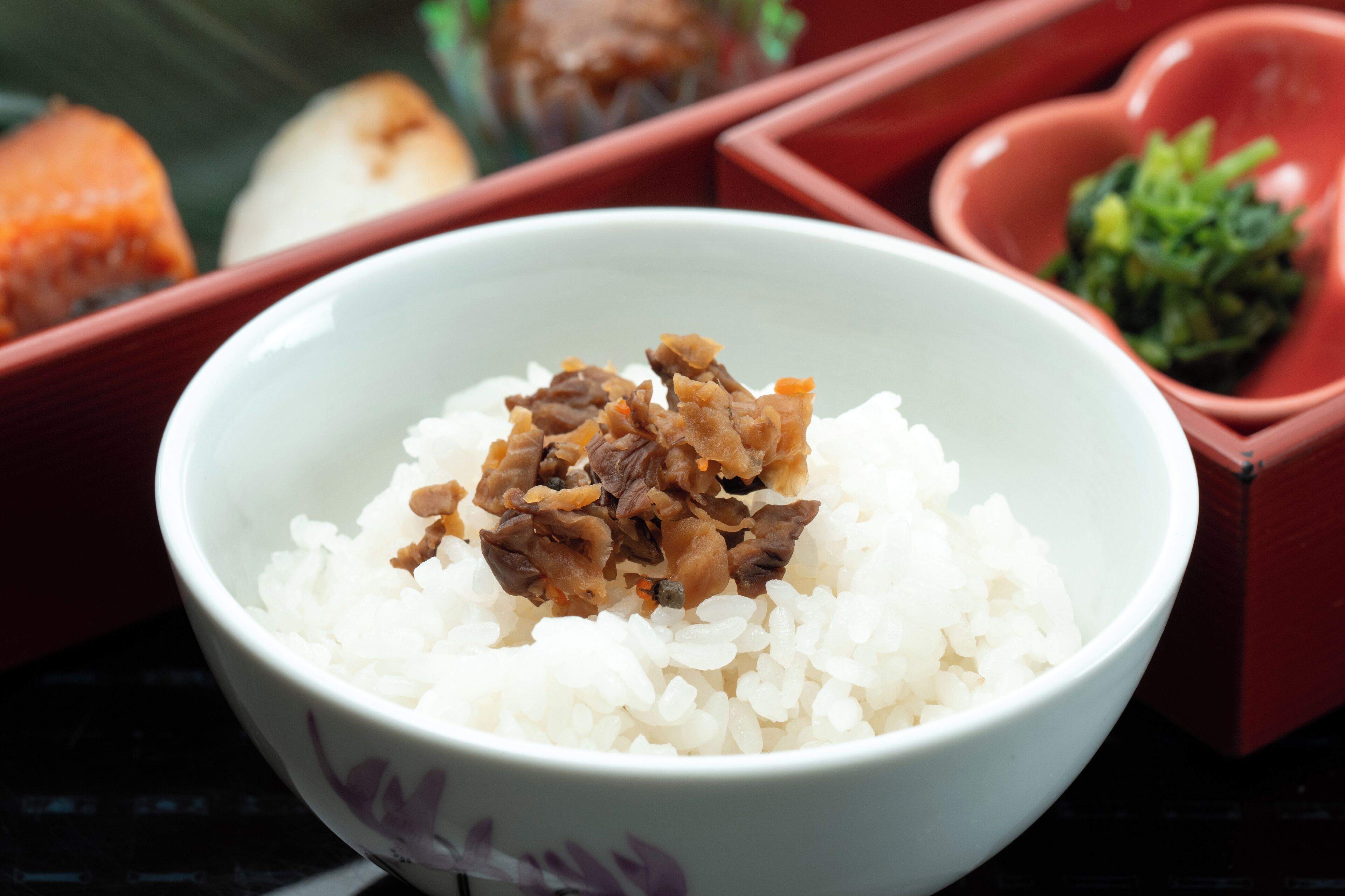 A bowl of white rice topped with brown ingredients, surrounded by red bowls containing other food items.