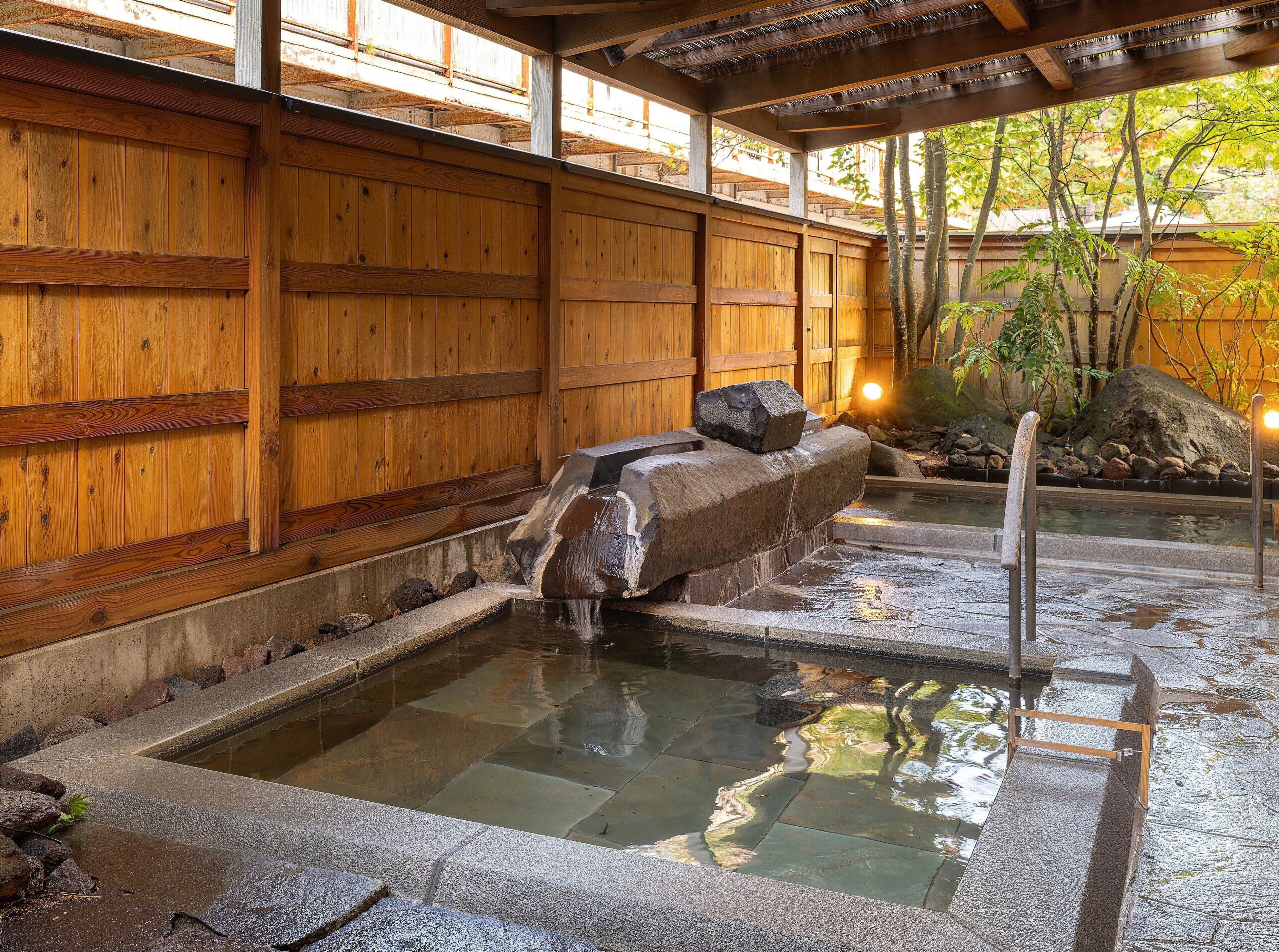 A traditional Japanese onsen with wooden walls, a stone hot spring, and a small waterfall.