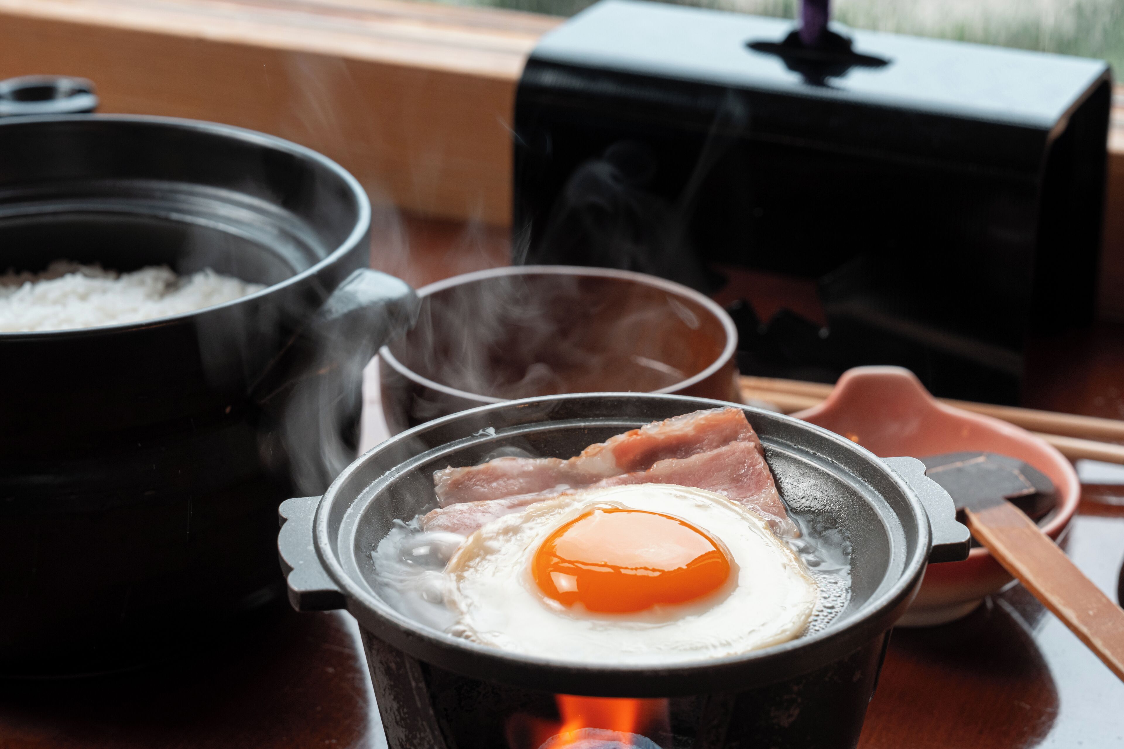 A hot pot with a cooked egg and bacon, a pot of rice, and a teapot on a table.