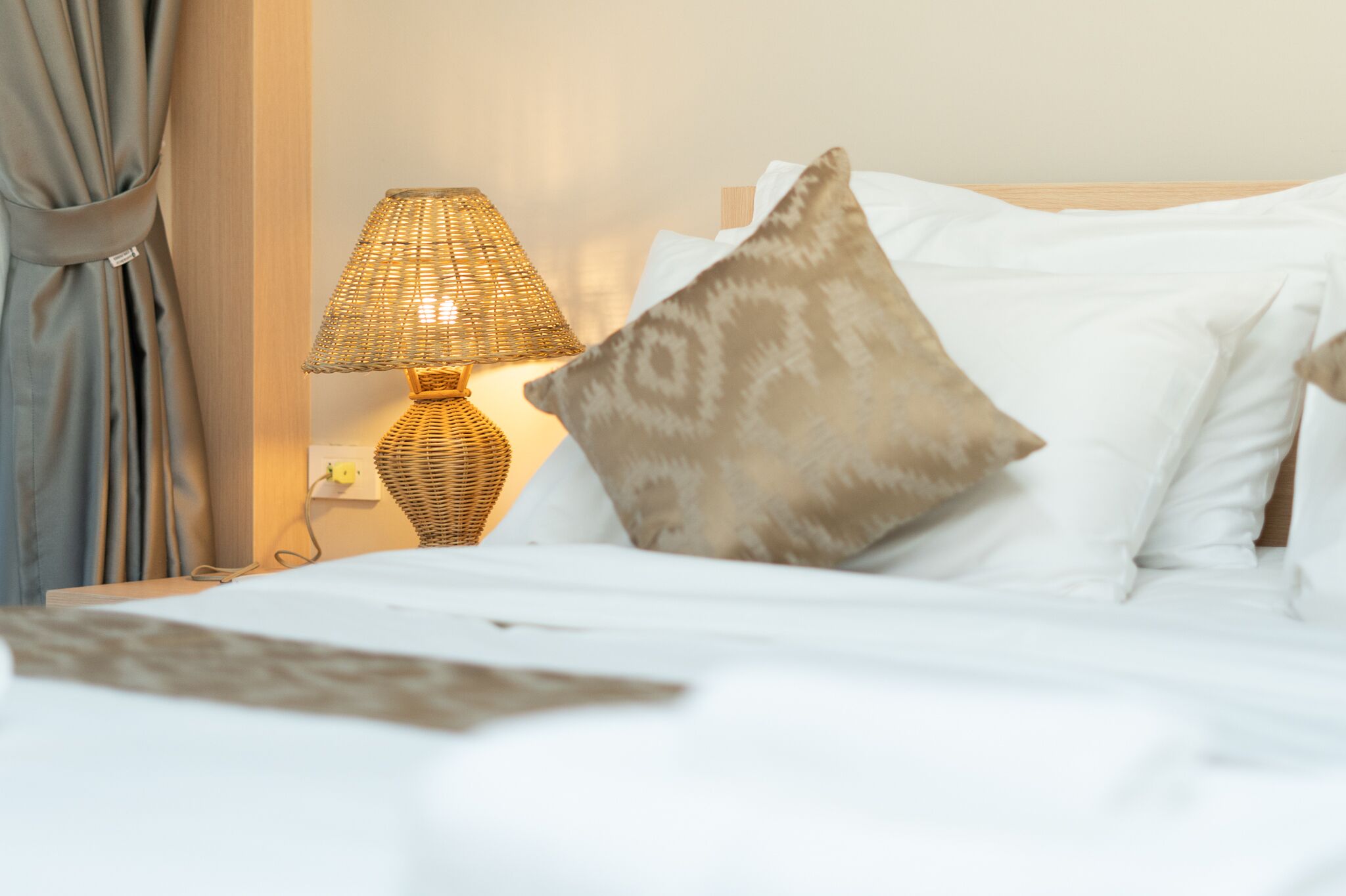 A neatly made bed with white linens, a patterned throw, and a woven table lamp on a bedside table.