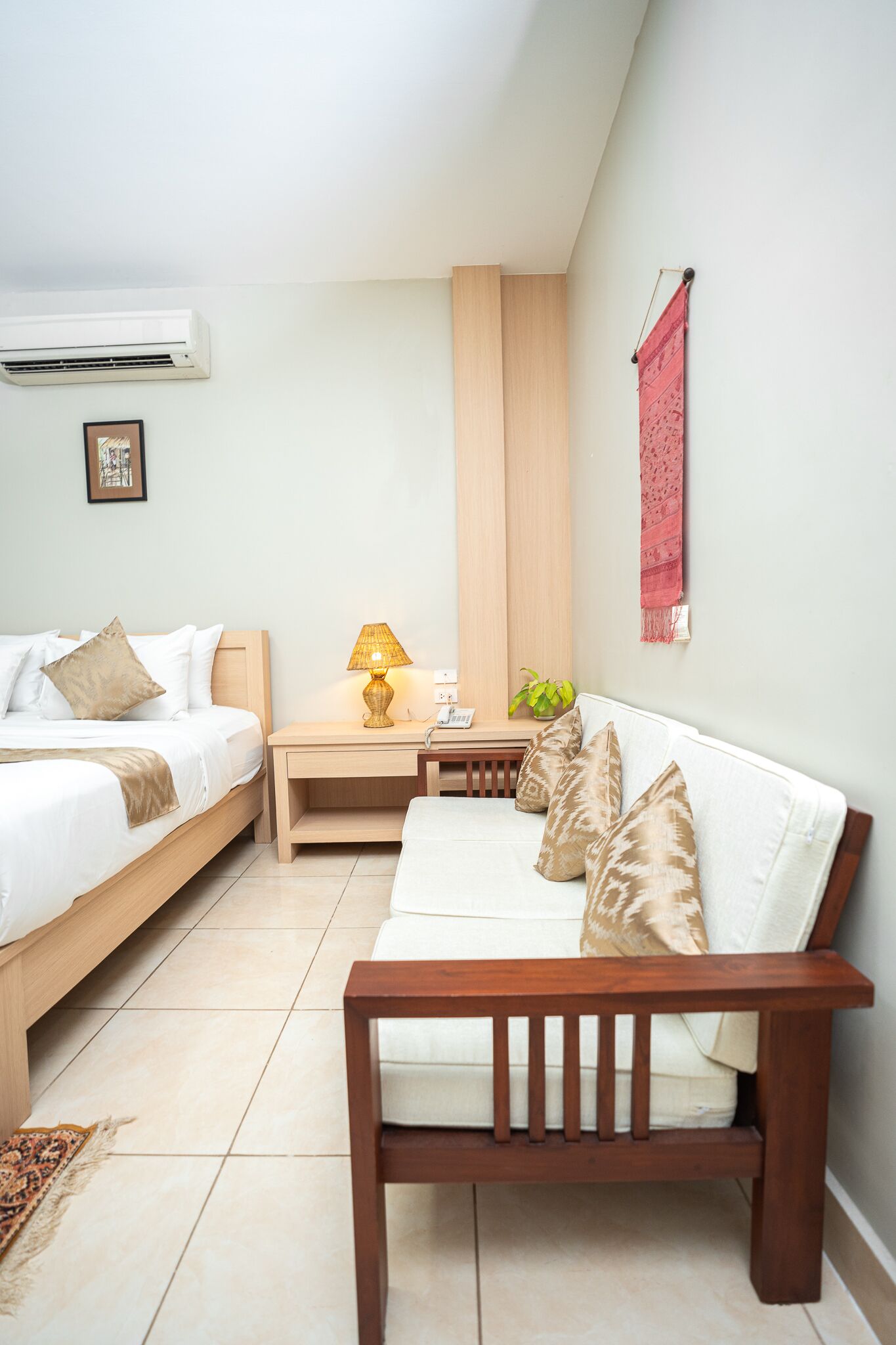 A bedroom with a bed, bedside table, lamp, and a red wall hanging.