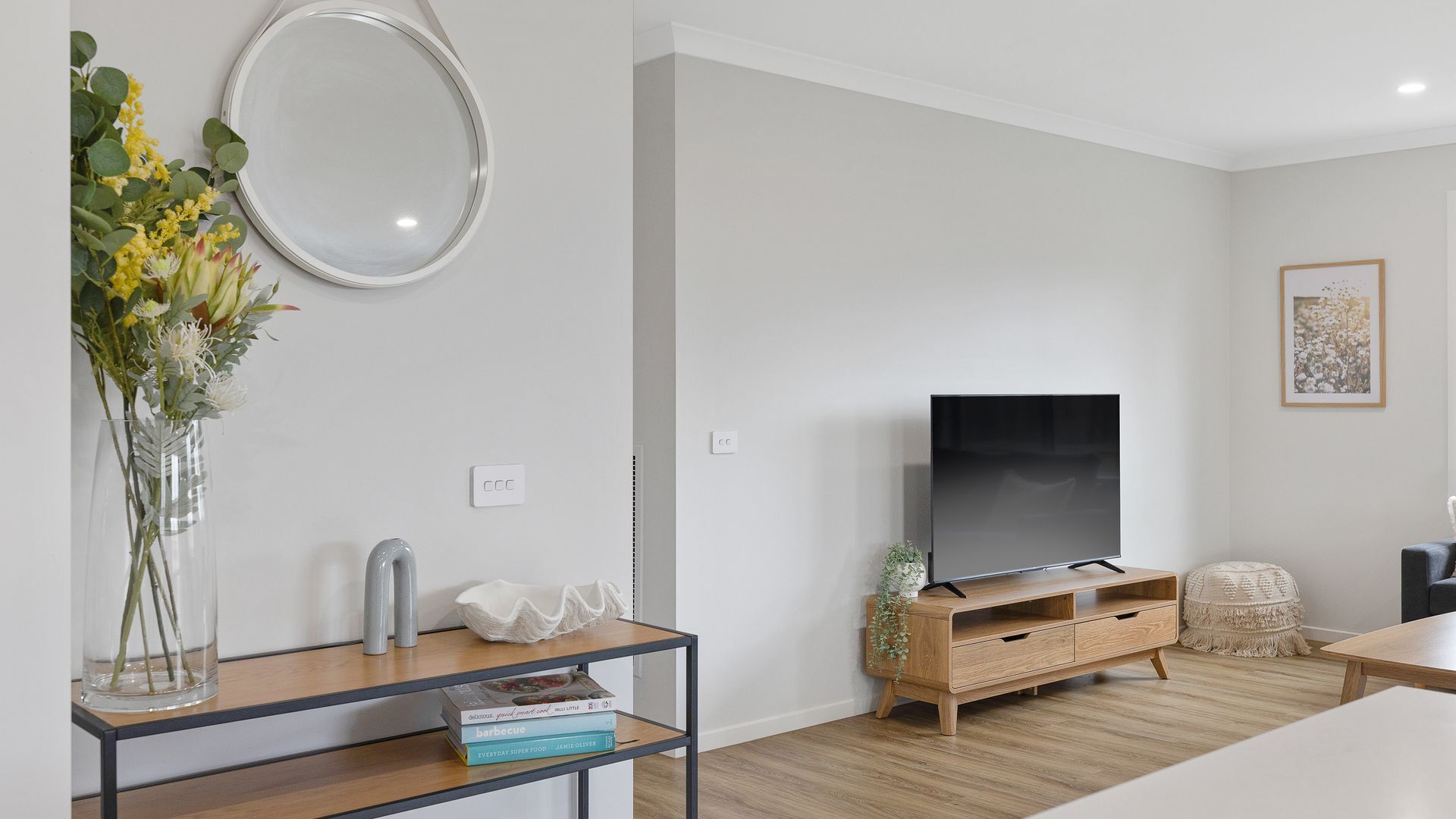 Een moderne woonkamer met een flatscreen televisie op een houten tv-meubel, een glazen vaas met bloemen op een bijzettafeltje en een ingelijste foto aan de muur.