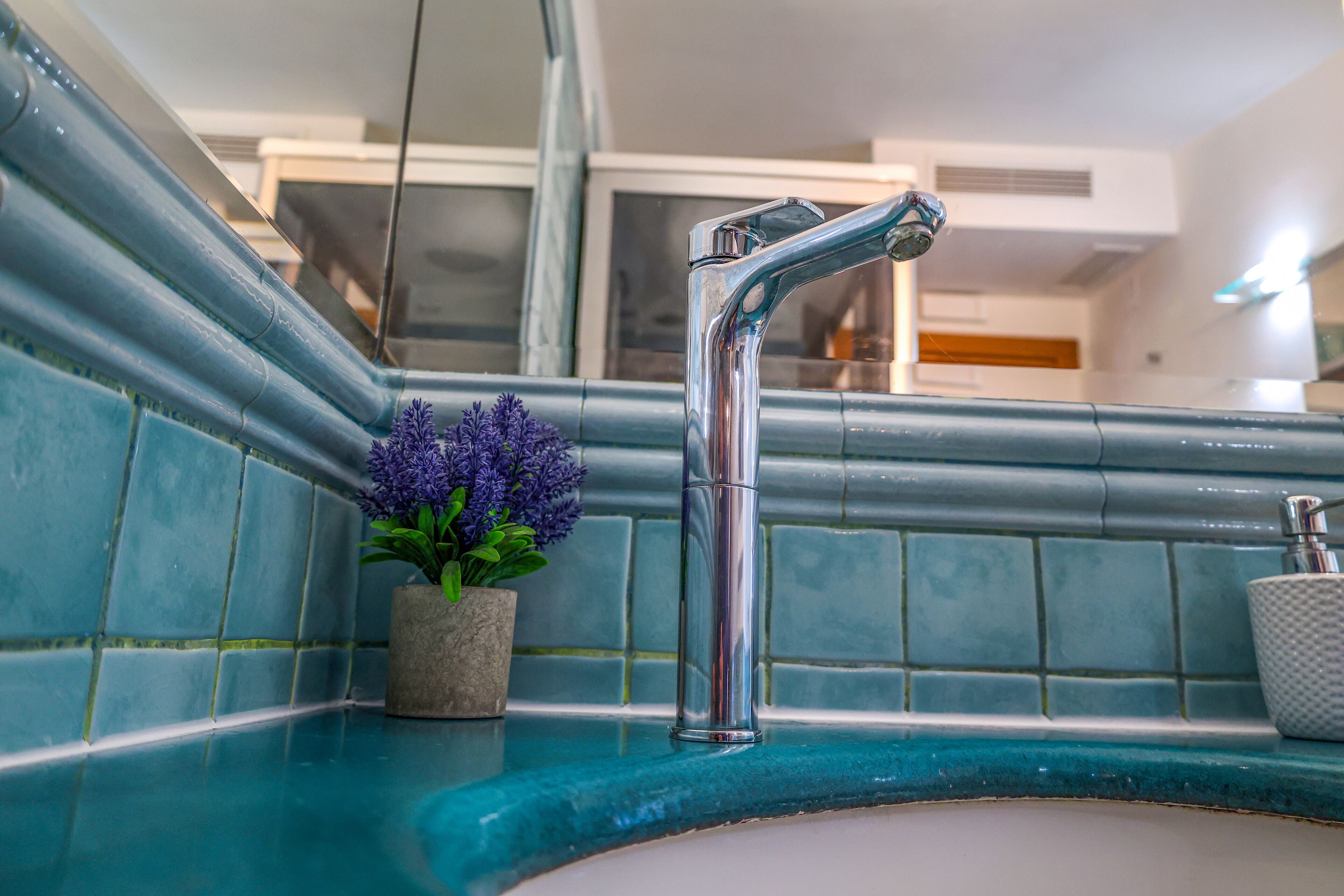 Une salle de bain dotée d’un robinet moderne, d’un pot de lavande et d’un distributeur de savon posés sur un comptoir recouvert de carreaux bleus.