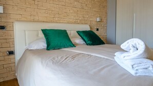 A neatly made bed with white linens and two green pillows, a white towel folded on the side, and a brick-patterned wall in the background. - villa Elisabetta (Pompei)