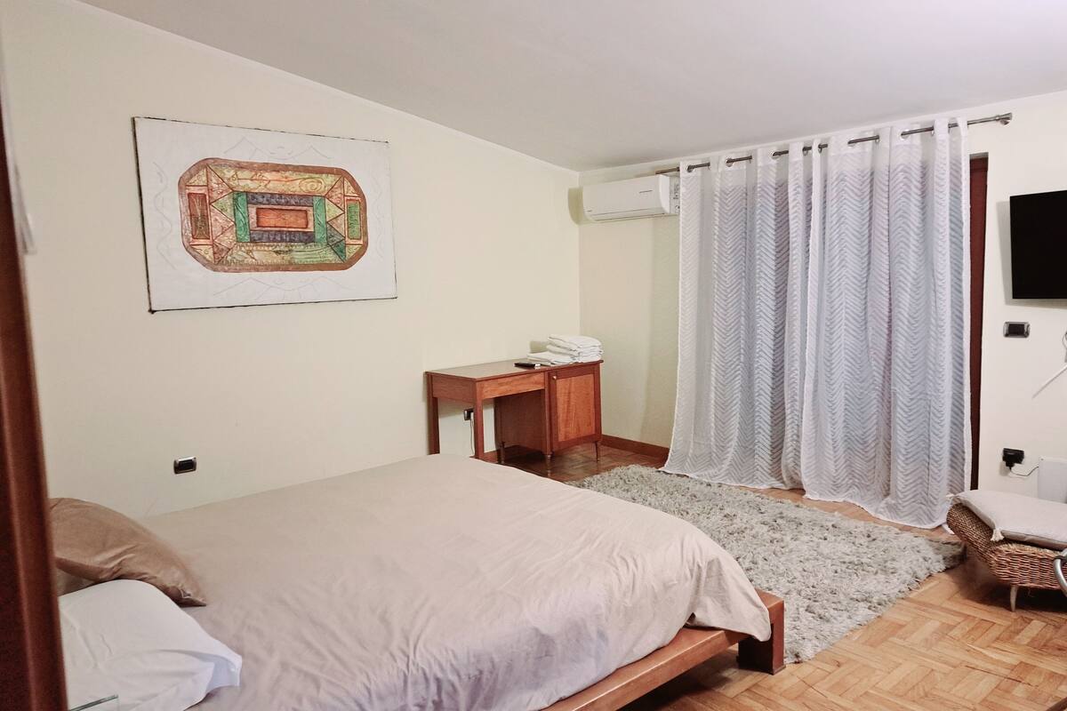 A bedroom with a bed, a desk, and a chair. There is a painting on the wall and a TV mounted on the wall.