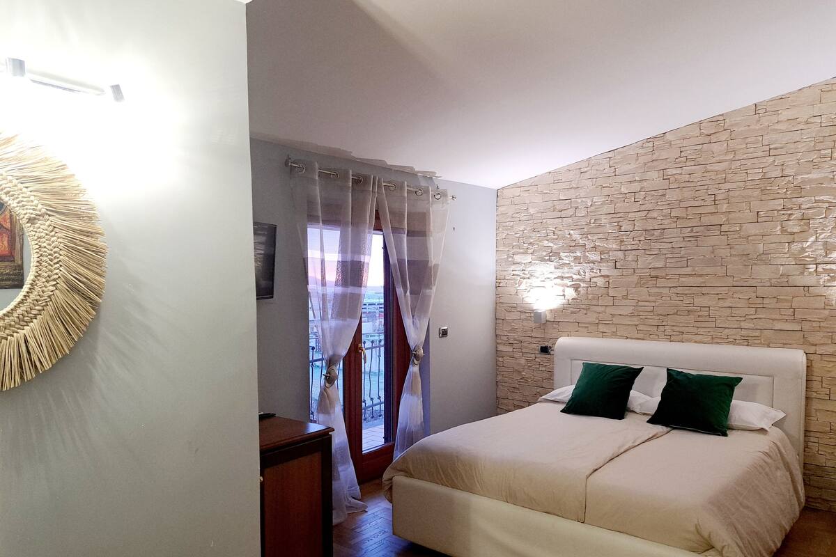 A bedroom with a bed, a wooden nightstand, a stone-textured wall, and a window with sheer curtains.