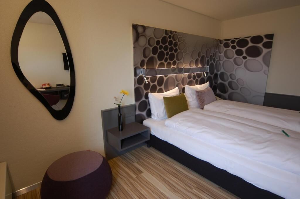 A modern hotel room with a large bed, a round mirror, a small side table, and a decorative wall panel.