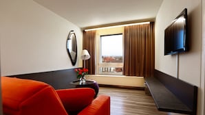 A hotel room with a modern design, featuring a sofa, a small table with a vase of flowers, a flat-screen TV mounted on the wall, and a window with curtains.