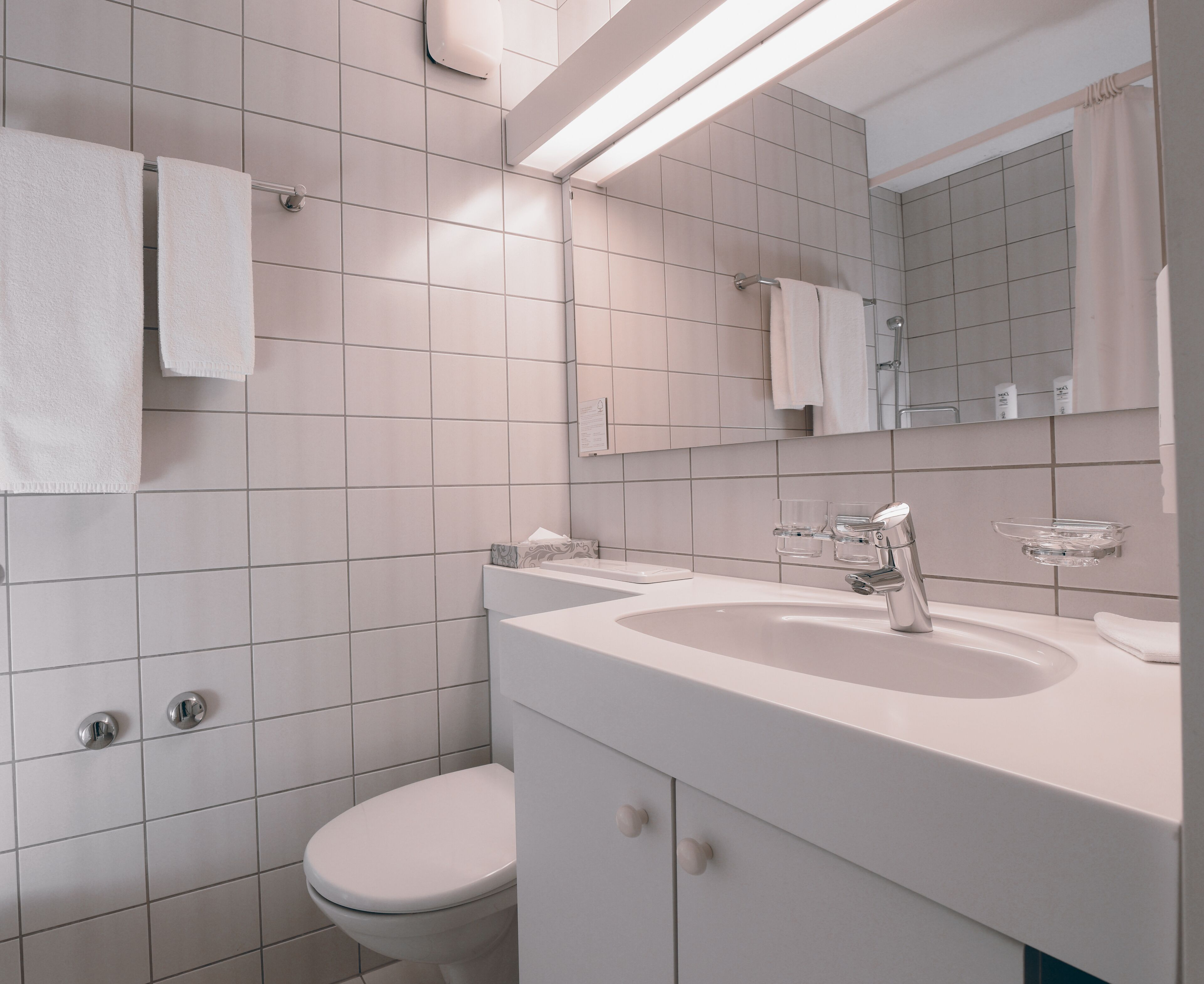 Un baño moderno con baldosas blancas, inodoro, lavabo con espejo y un perchero para toallas.