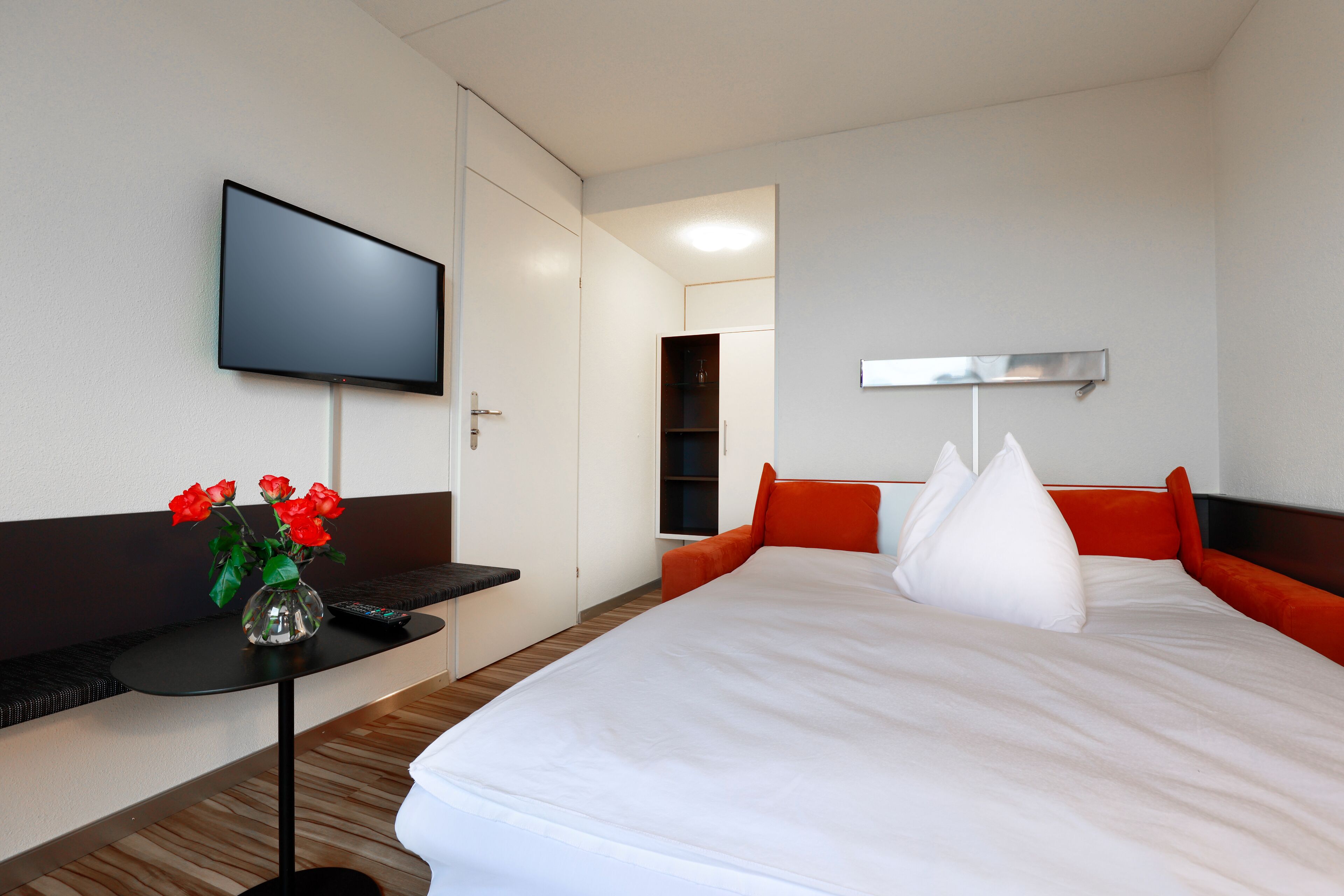 A hotel room with a bed, a TV mounted on the wall, a small table with a vase of flowers, and a wardrobe.