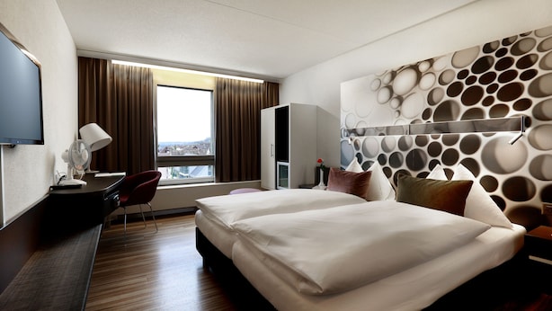 A hotel room with a bed, a desk, a chair, a TV, and a window with curtains.