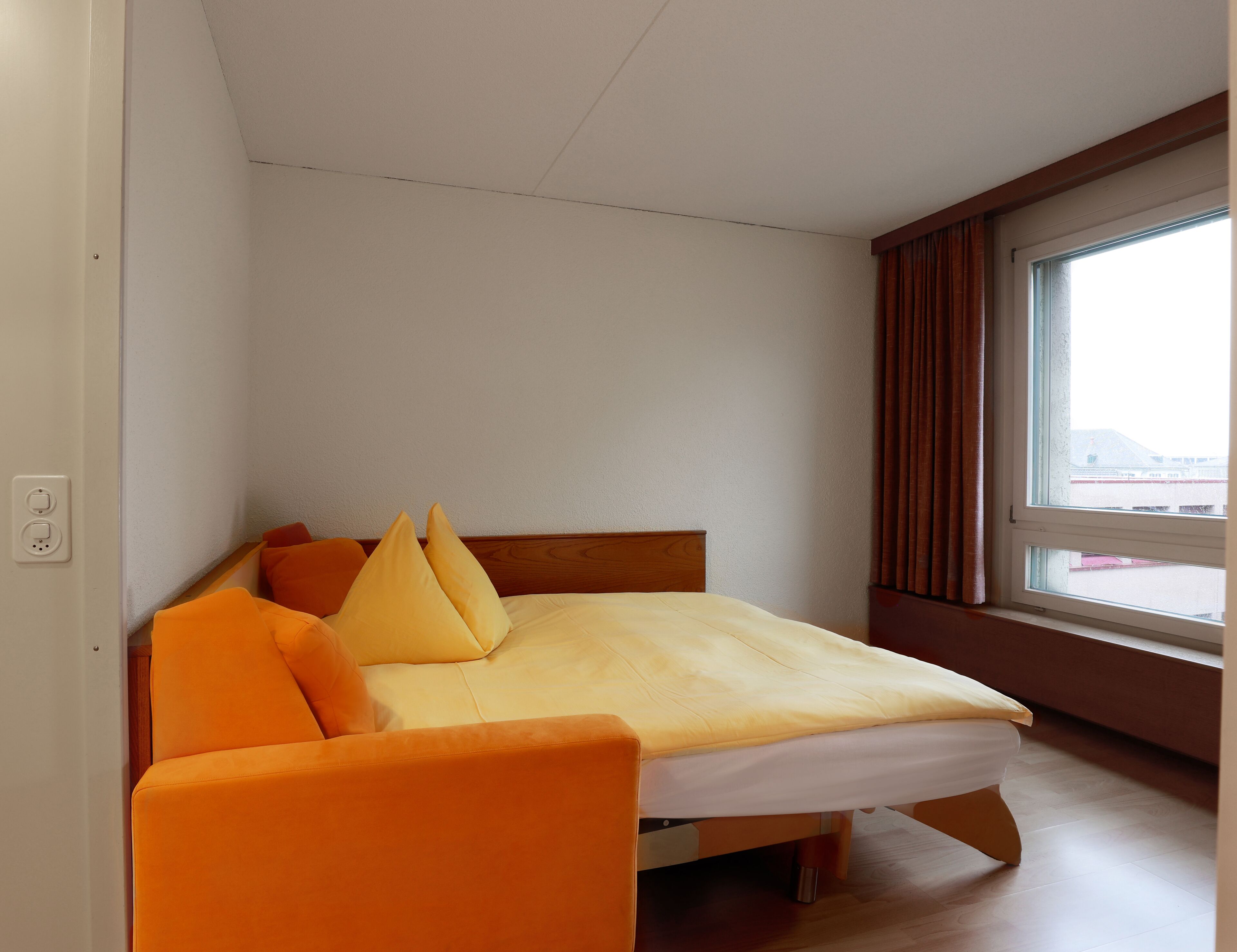 Una cama individual con estructura de madera y colchón amarillo, almohadas naranjas y cabecera de madera. Se ve una ventana con cortinas en el lado derecho.