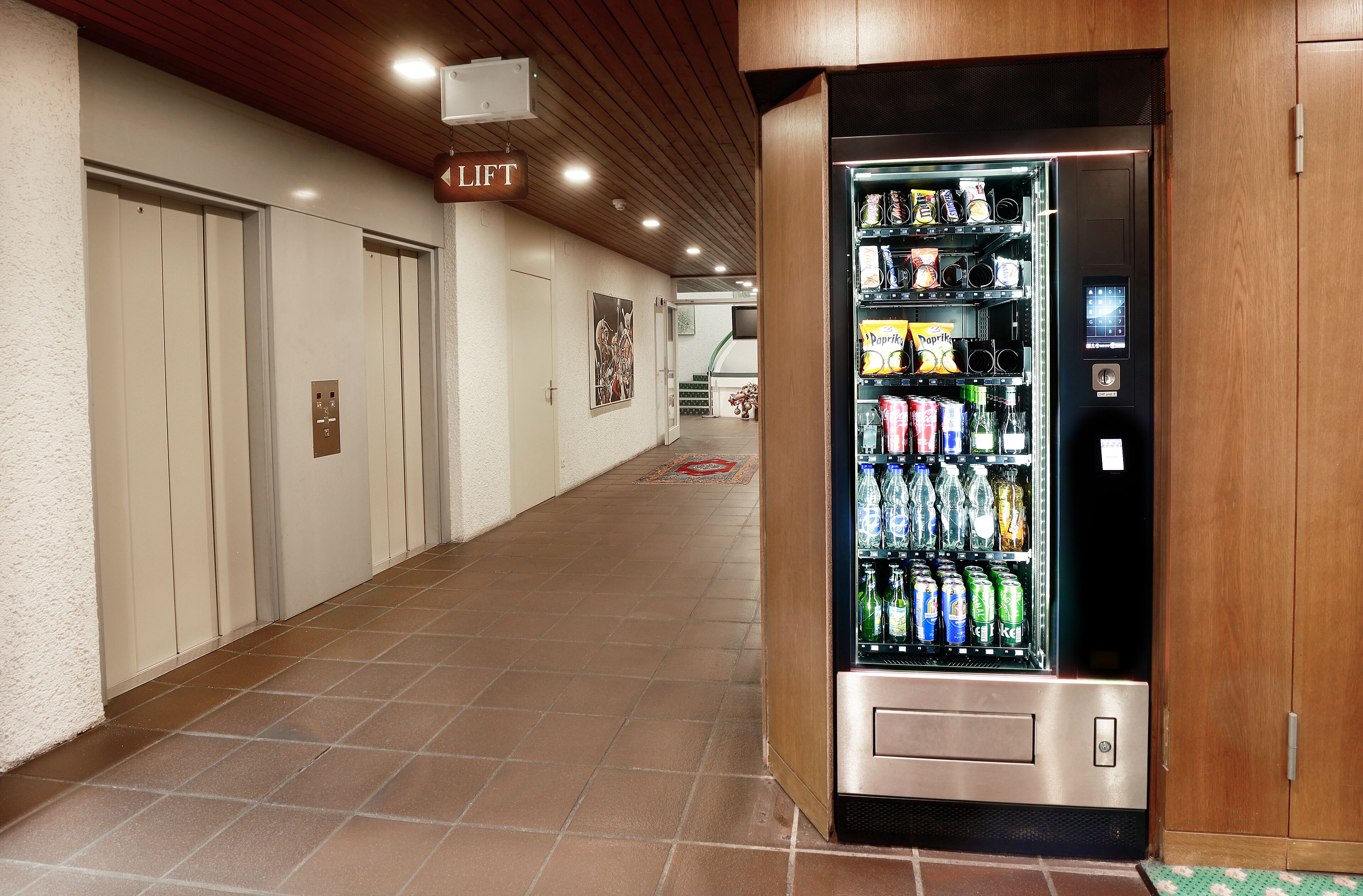 Een gang met een automaat vol drankjes, een lift en een bord met 'Lift' erboven.