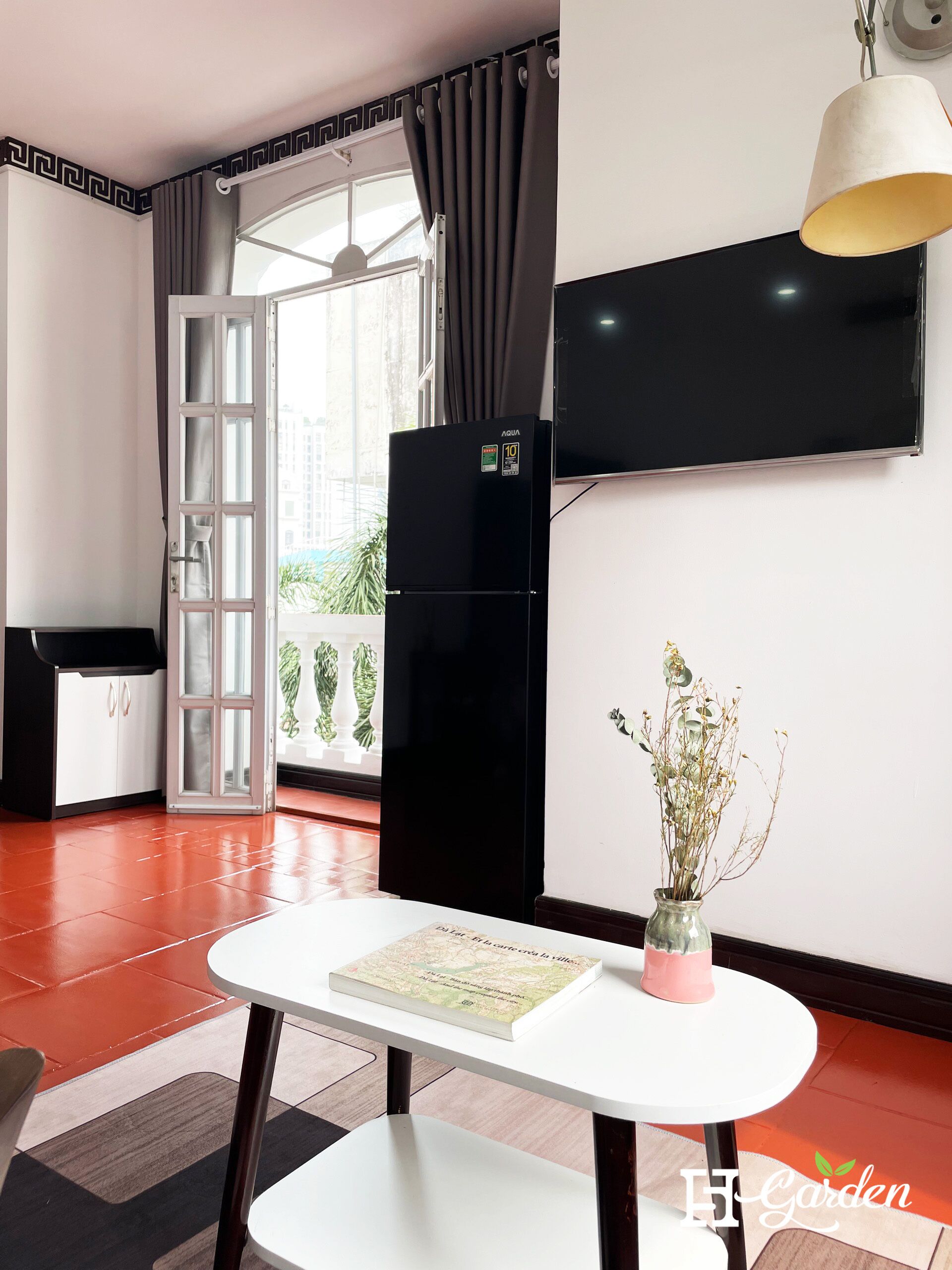 Un soggiorno moderno con un frigorifero nero, una TV a schermo piatto montata a parete, un tavolo da pranzo con un libro e un vaso di fiori secchi, e vista su un balcone con piante.